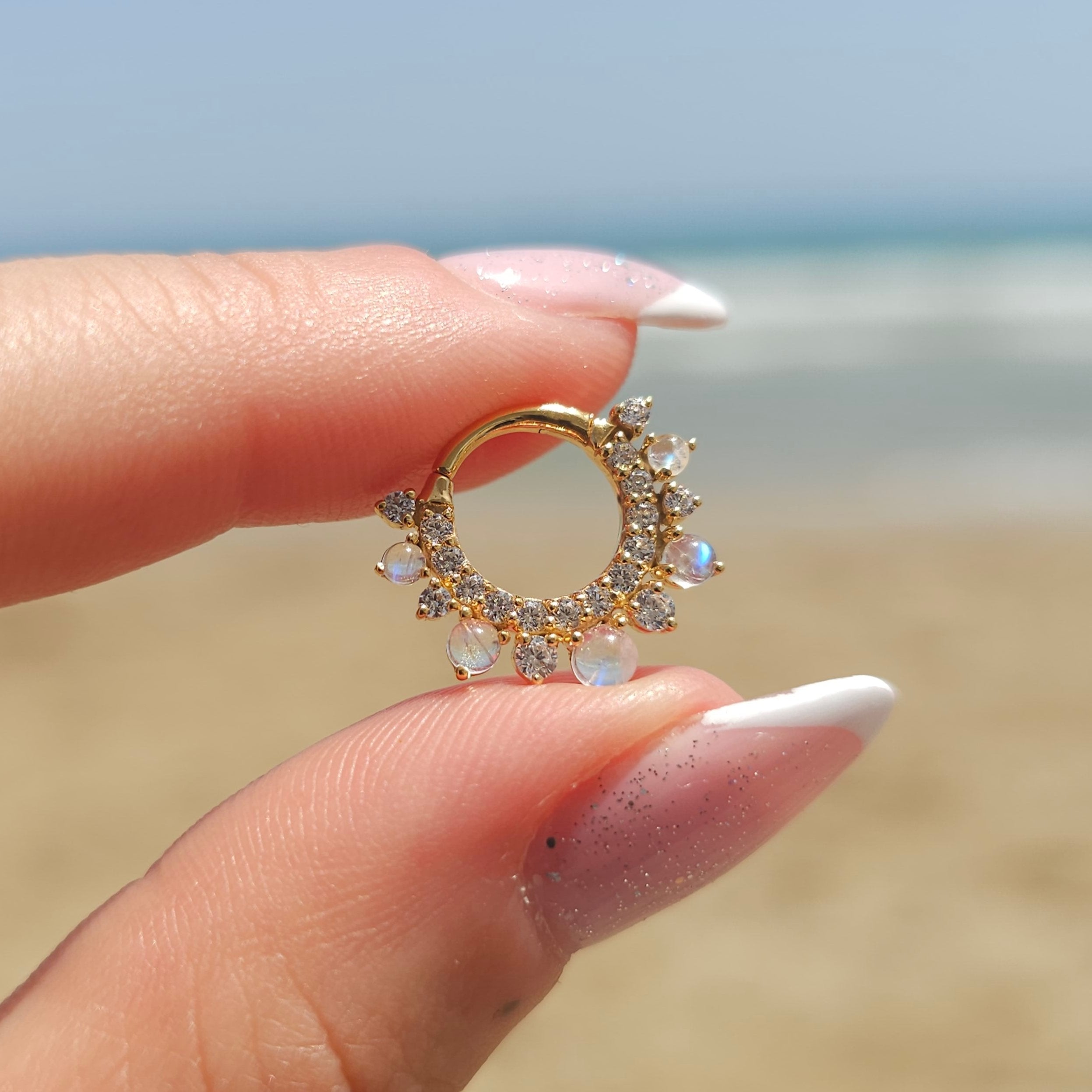 A manicured hand holds the CHLUXE • CALYPSO Moonstone Daith Hoop by Crystal Heaven London, a 14K Yellow Gold limited edition piece adorned with rainbow moonstones, against a blurred beach background.