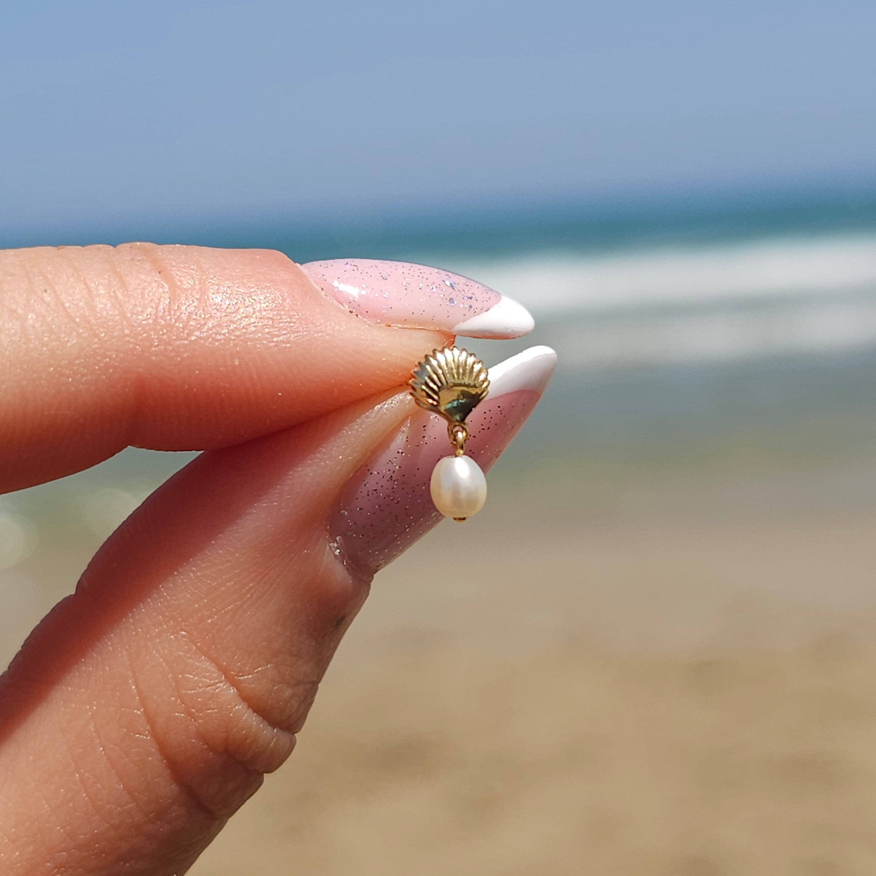 In a softly blurred beach and ocean backdrop, a hand delicately holds the CHLUXE • MILOS Cartilage Stud by Crystal Heaven London—a Limited Edition 14K Yellow Gold seashell-shaped earring with a pearl that exudes elegance and seaside charm.