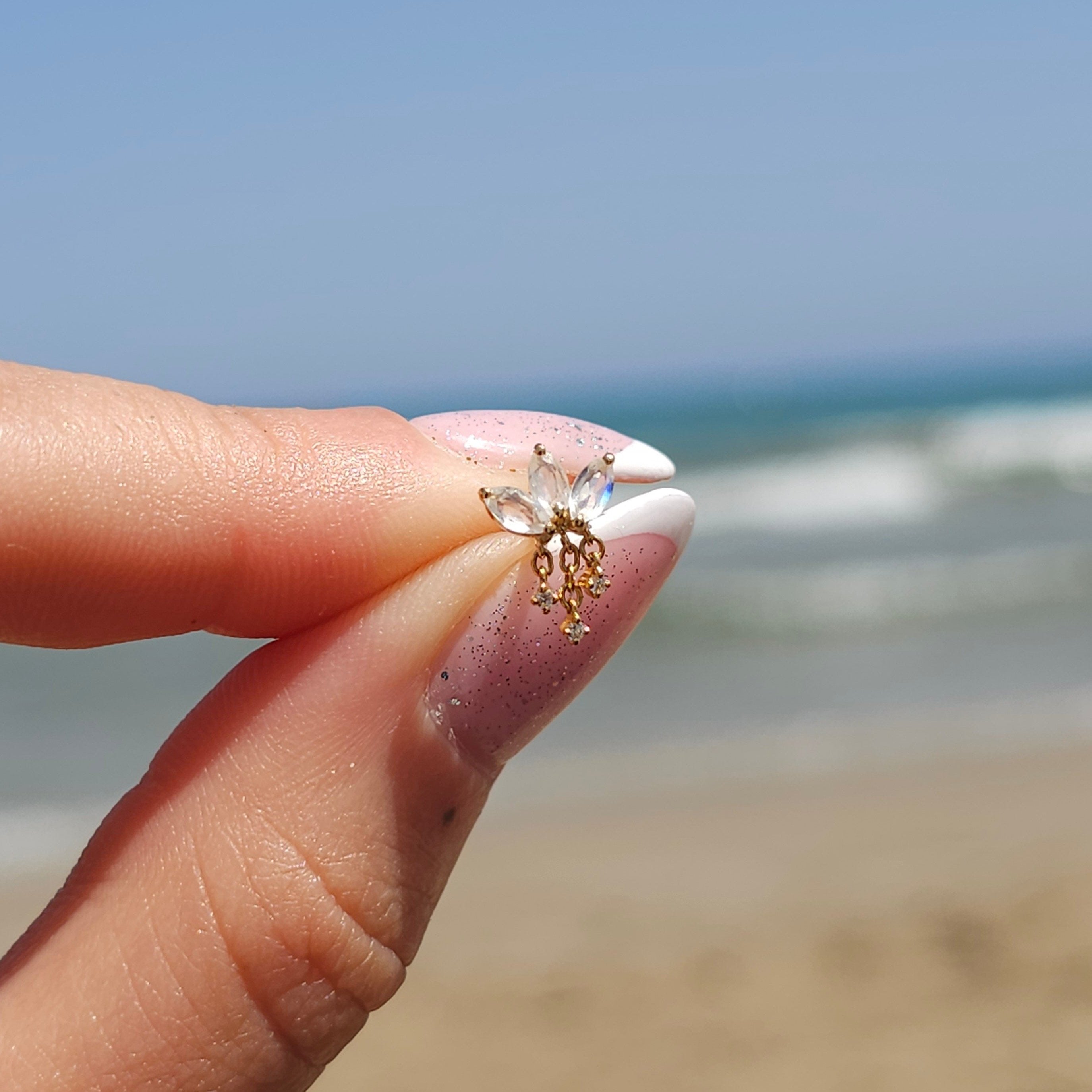A manicured hand gracefully holds a CHLUXE • LEILANI Chain Droplet Cartilage Stud, a Limited Edition piece by Crystal Heaven London crafted from 14k Yellow Gold, set against a tranquil beach and ocean background.