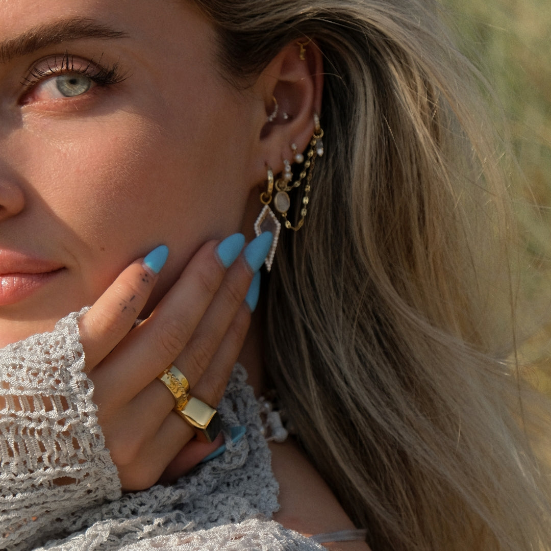 A person with long blonde hair and blue eyes wears gold jewelry from the Nerida Collection, featuring the SIRENA Moonstone Charm by Crystal Heaven London. With blue nail polish, they rest their hand on their cheek, partially covering their face.