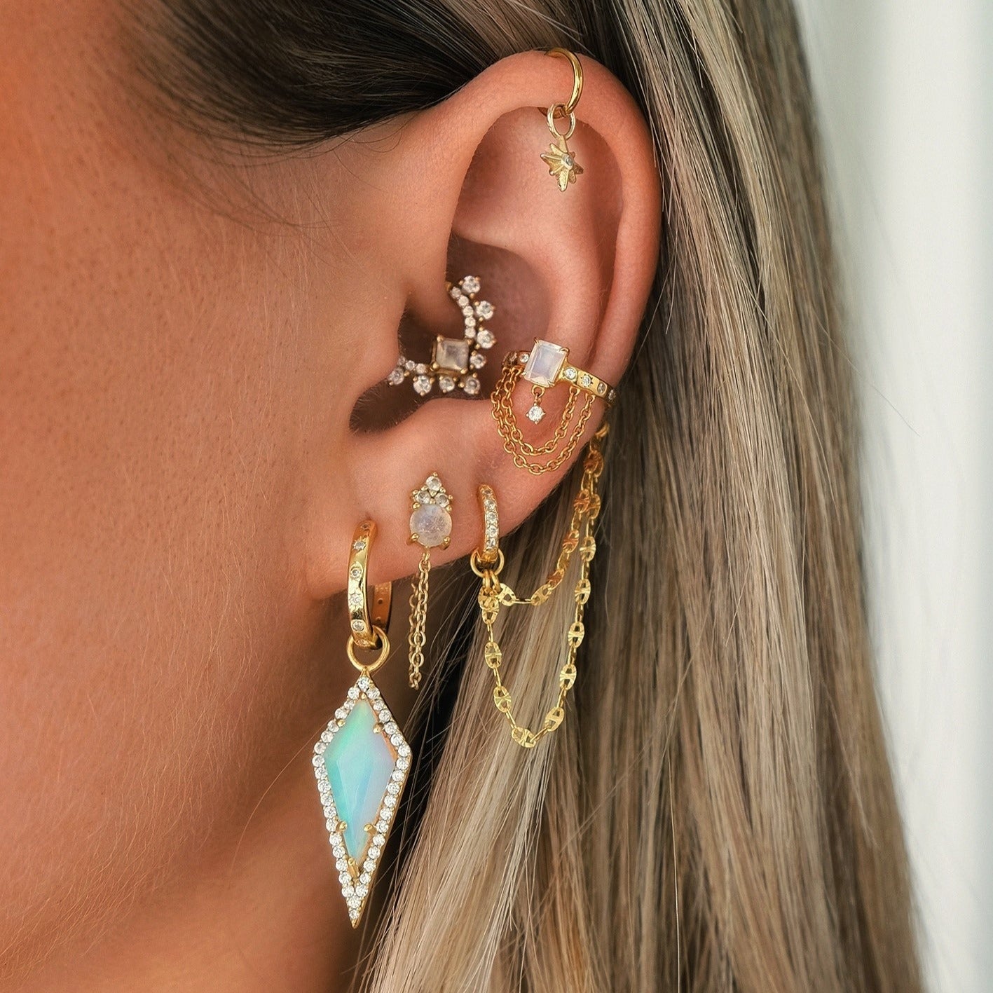 A close-up of an ear showcases gold piercings with chains, gemstones, intricate designs, and a large diamond-shaped turquoise earring. The look is complemented by Crystal Heaven London's HAVANA Slim Starburst Huggie Hoops and delicate starburst-detailed earring charms.