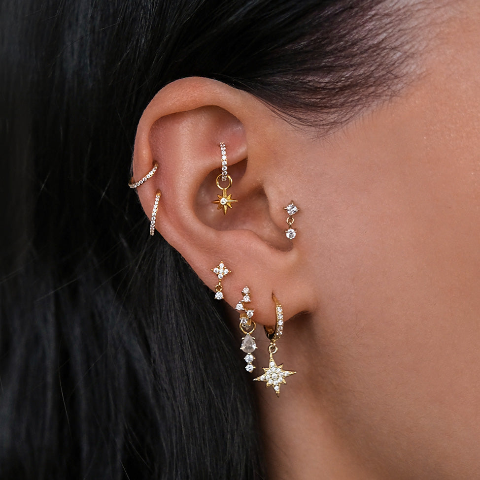 A beautifully adorned ear with multiple piercings showcases a CHLUXE Original Design by Crystal Heaven London, featuring the VENUS Cartilage Stud in 14K Yellow Gold with hoops, studs, dangling designs, small gems, and enchanting starburst shapes.