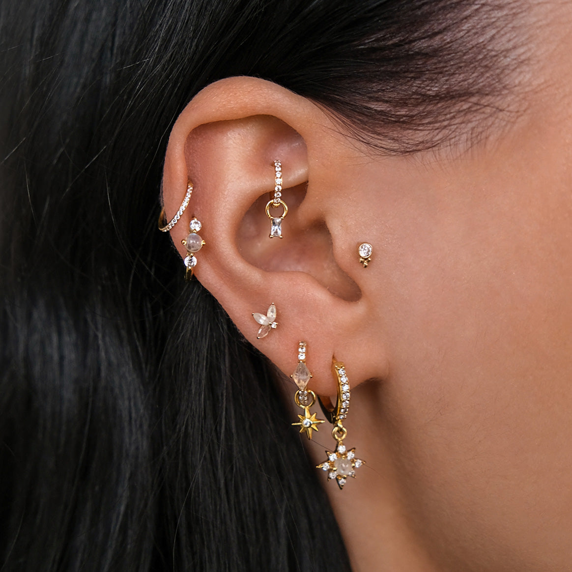 Close-up of an ear with multiple piercings, including the CHLUXE • KAMALA Cartilage Stud in 14K Yellow Gold by Crystal Heaven London, alongside hoops and studs featuring star, butterfly, and lotus designs accented with Moonstone gemstones.