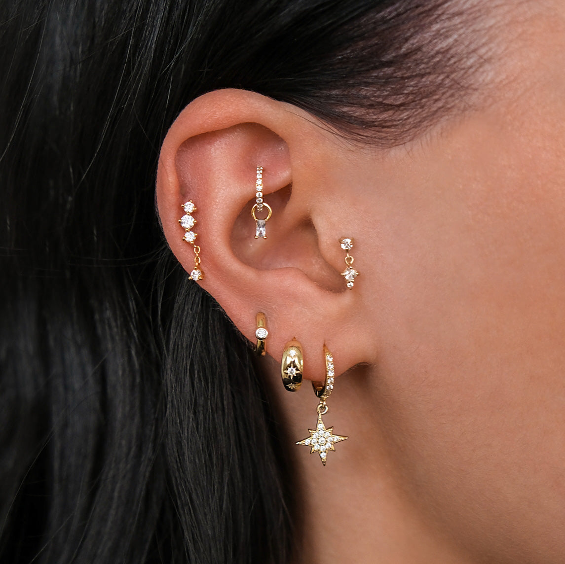 Close-up of a person's ear featuring CHLUXE • LUCID Droplet Cartilage Stud in 14K Gold by Crystal Heaven London, alongside hoops, studs, and a star dangle earring. The dark glossy hair enhances the LUCID elegance.