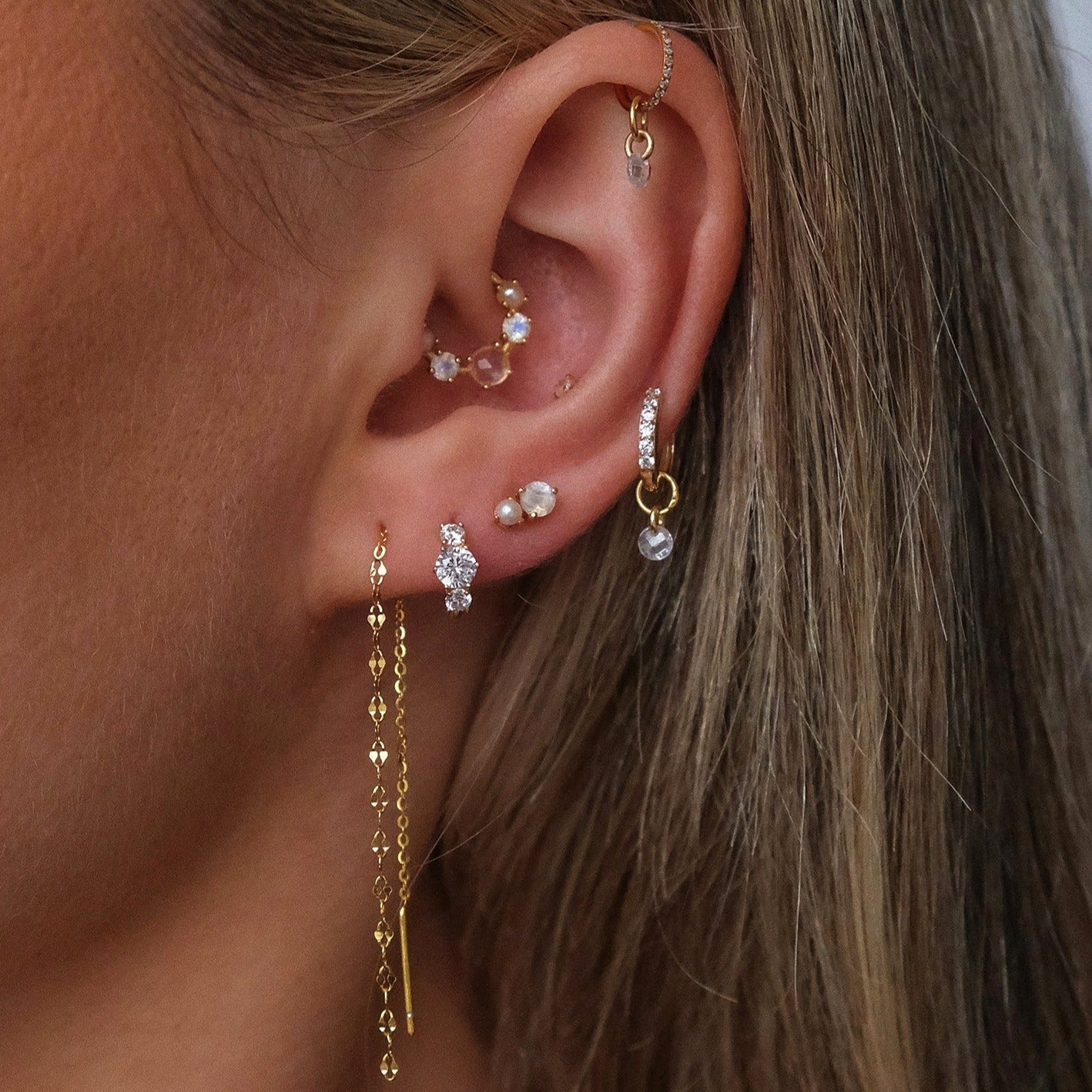Close-up of an ear with multiple piercings showcasing 14K solid yellow gold earrings, including small studs, hoops, a CHLUXE Stacked Crystal Huggie Hoop by Crystal Heaven London, and one long dangling chain earring.