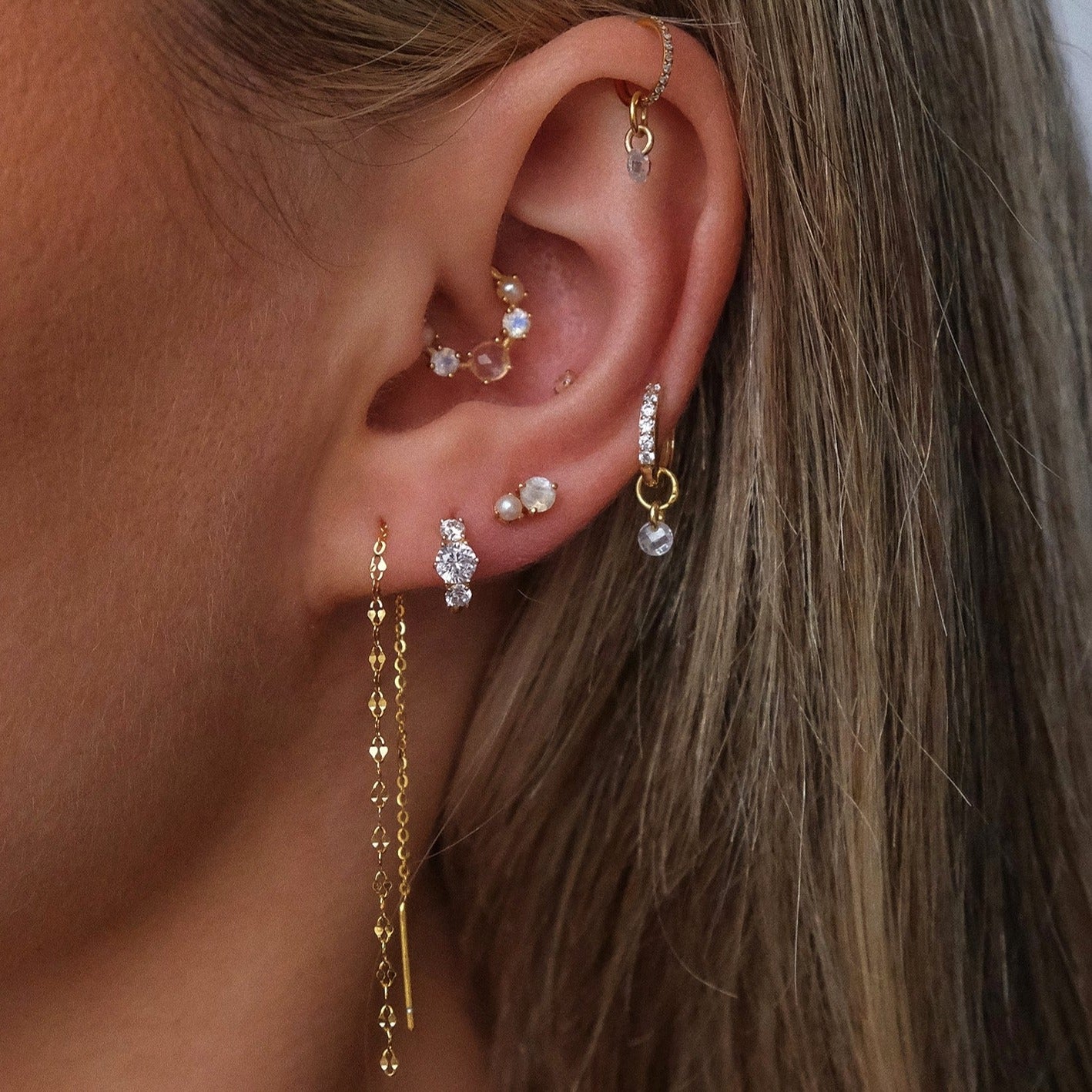 A person's ear features Crystal Heaven London's CHLUXE • OPHELIA 14K gold cartilage stud, hoops with pearls and gemstones, and a long dangling chain inspired by Ophelia's ethereal style.