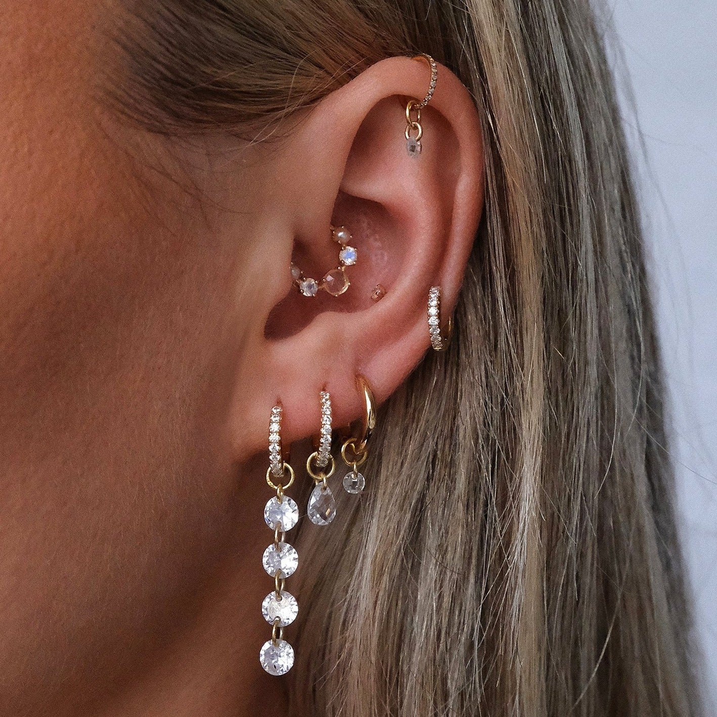 A close-up highlights a person's ear adorned with multiple gold earrings, including Crystal Heaven London's ALLURE Cascading Crystal Huggie Hoops, showcasing glistening pavé, crystals, and small hoops.