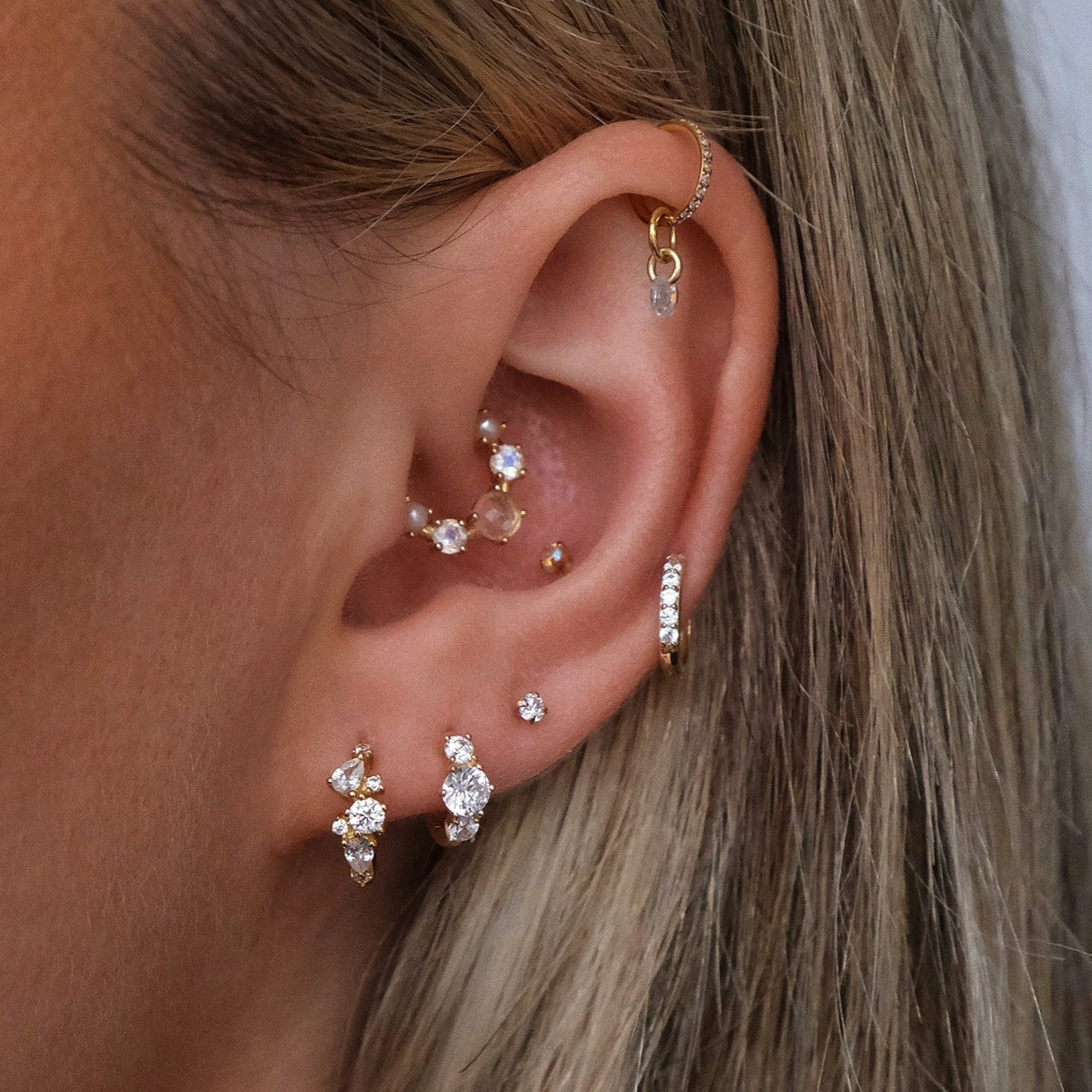 A close-up shot features multiple piercings adorned with Crystal Heaven London's MARGOT Scattered Gem Huggie Hoops and other sparkling sterling silver earrings, some with cubic zirconia stones, arranged along the ear's helix, tragus, and lobe.