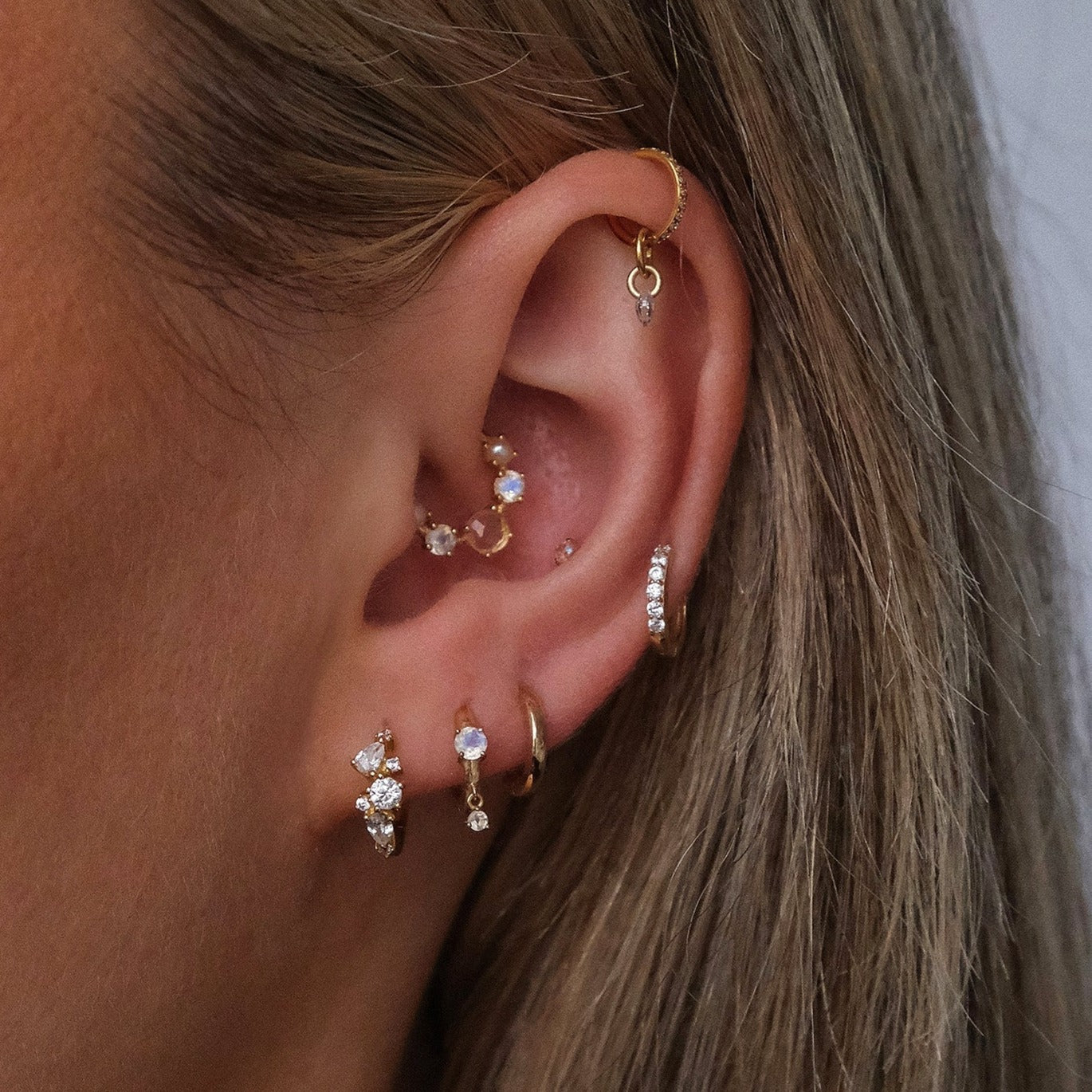 Close-up of an ear with multiple piercings featuring CHLUXE Moonstone Droplet Huggie Hoop - 14K Yellow Gold by Crystal Heaven London, adorned with delicate moonstones.