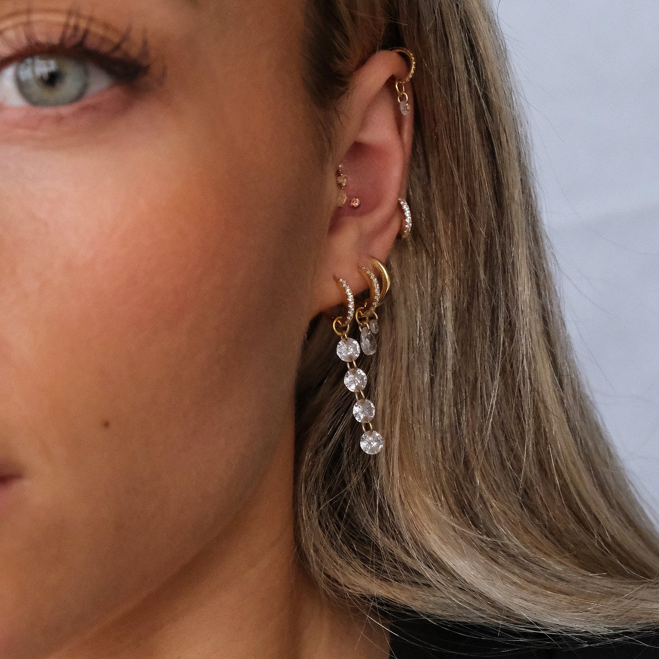 A close-up of a person's ear displays multiple piercings with gold earrings, including the CHLUXE Crystal Huggie Hoop by Crystal Heaven London, encrusted with clear beads and cubic zirconia, all set in 14K yellow gold.