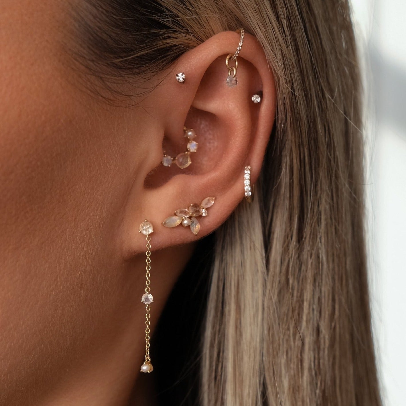 A close-up of a person's ear reveals multiple piercings featuring an array of earrings, including OPHELIA Gemstone Waterfall Studs by Crystal Heaven London, as well as hoops and dangly styles adorned with rose-cut white topaz and radiant rainbow moonstone.