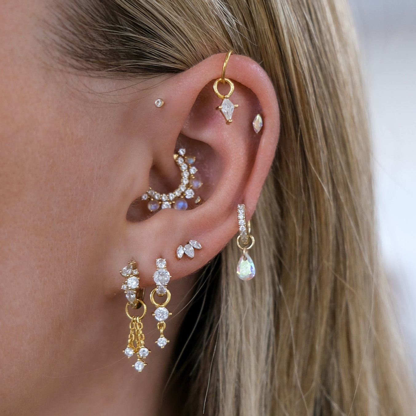 A close-up of an ear adorned with gold and diamond earrings, including hoops, studs, and dangles featuring Crystal Heaven London's VOGUE Droplet Charm with cubic zirconia highlights, showcasing intricate piercings.