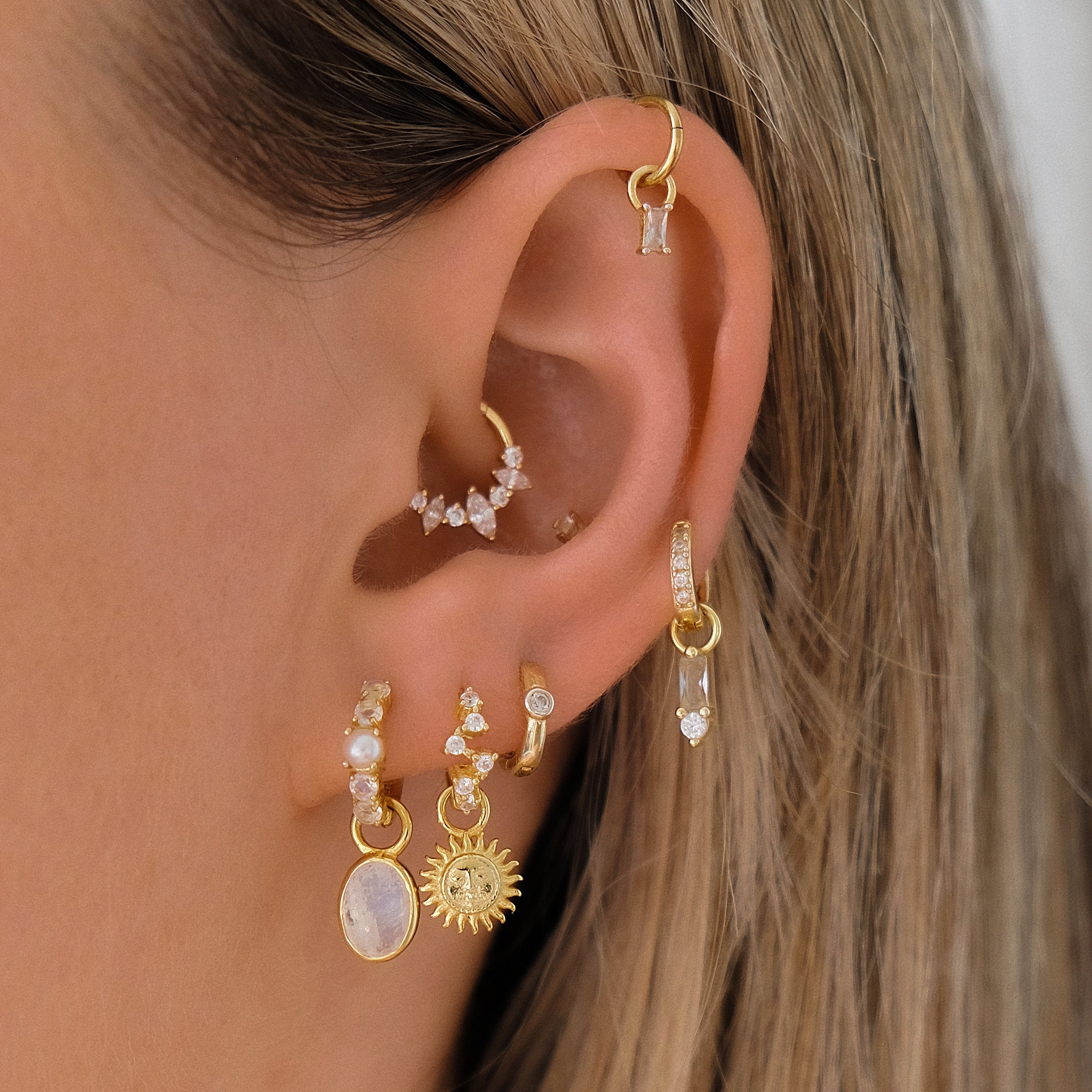 A close-up shows an ear with various gold earrings, including studs and hoops, from Crystal Heaven London. The PERLA Moonstone & Pearl Huggie Hoops add a mystical shimmer with freshwater pearls, crystals, charms, and rainbow moonstone.