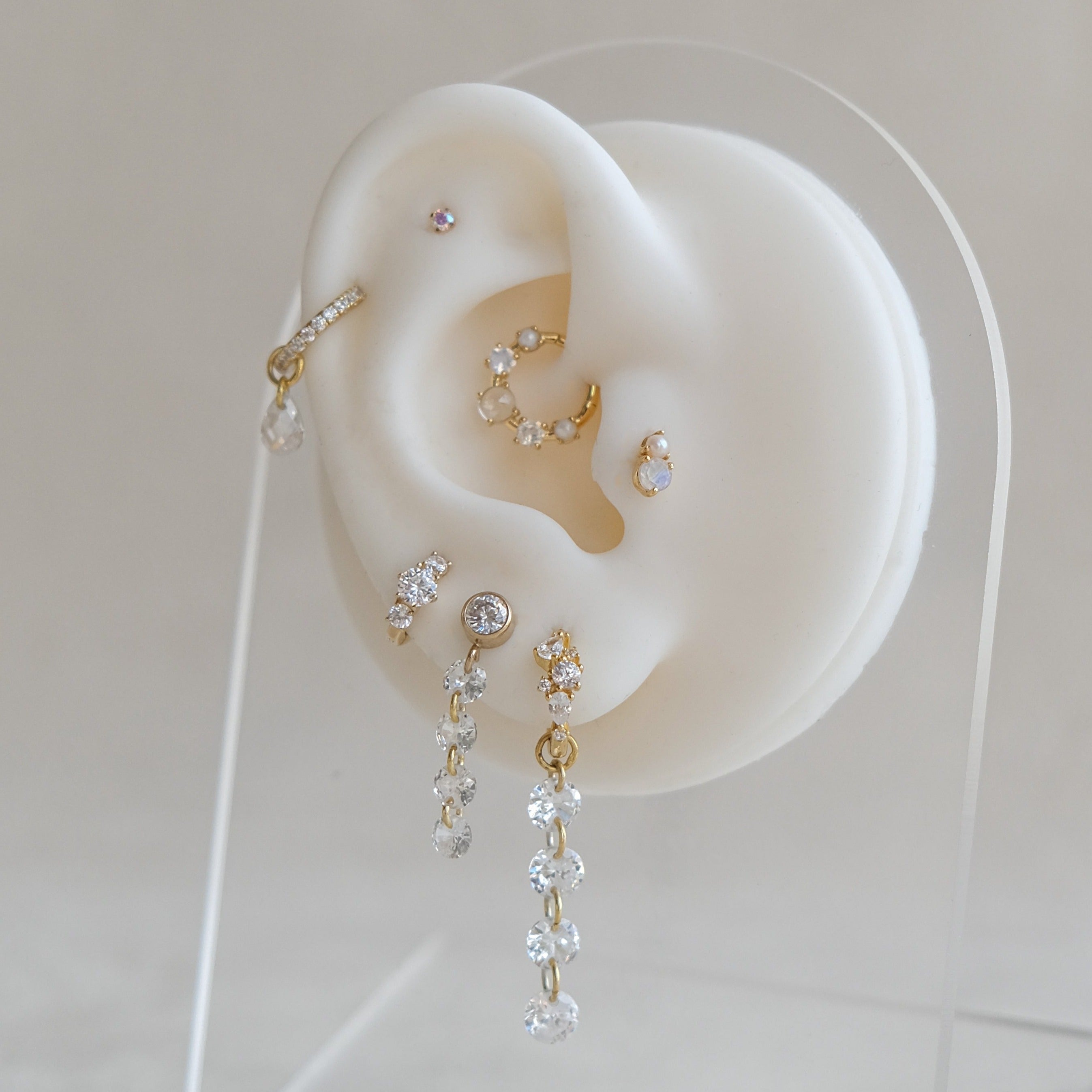 A plastic ear model displays various piercings featuring the Crystal Heaven London CHLUXE Stacked Crystal Huggie Hoops, crafted in 14K solid yellow gold with diamond and AAAA cubic zirconia earrings, elegantly set on a clear stand.