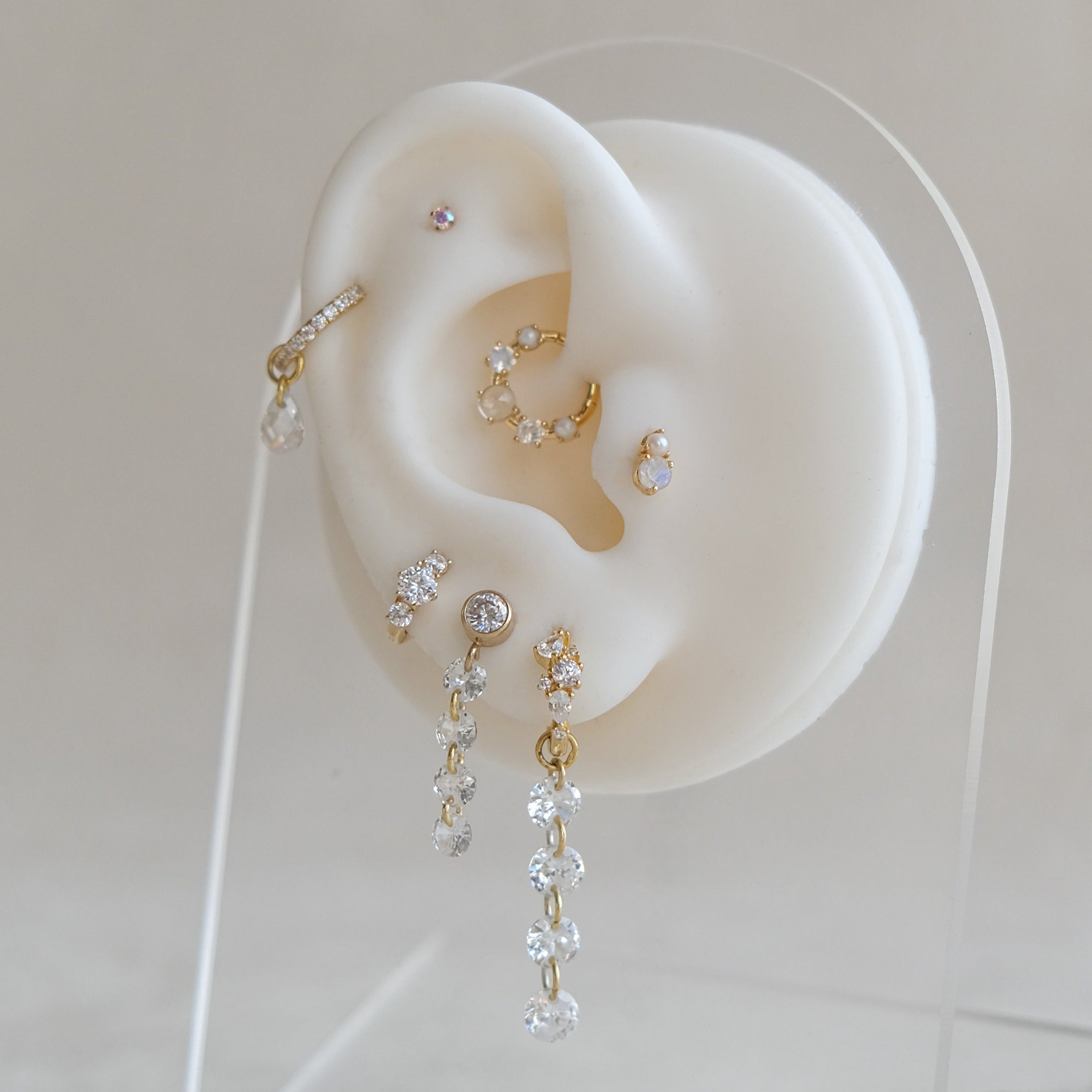The white ear-shaped earring holder showcases eye-catching gold and sterling silver earrings, including Crystal Heaven London's MARGOT Scattered Gem Huggie Hoops, sparkling with cubic zirconia and other dazzle.