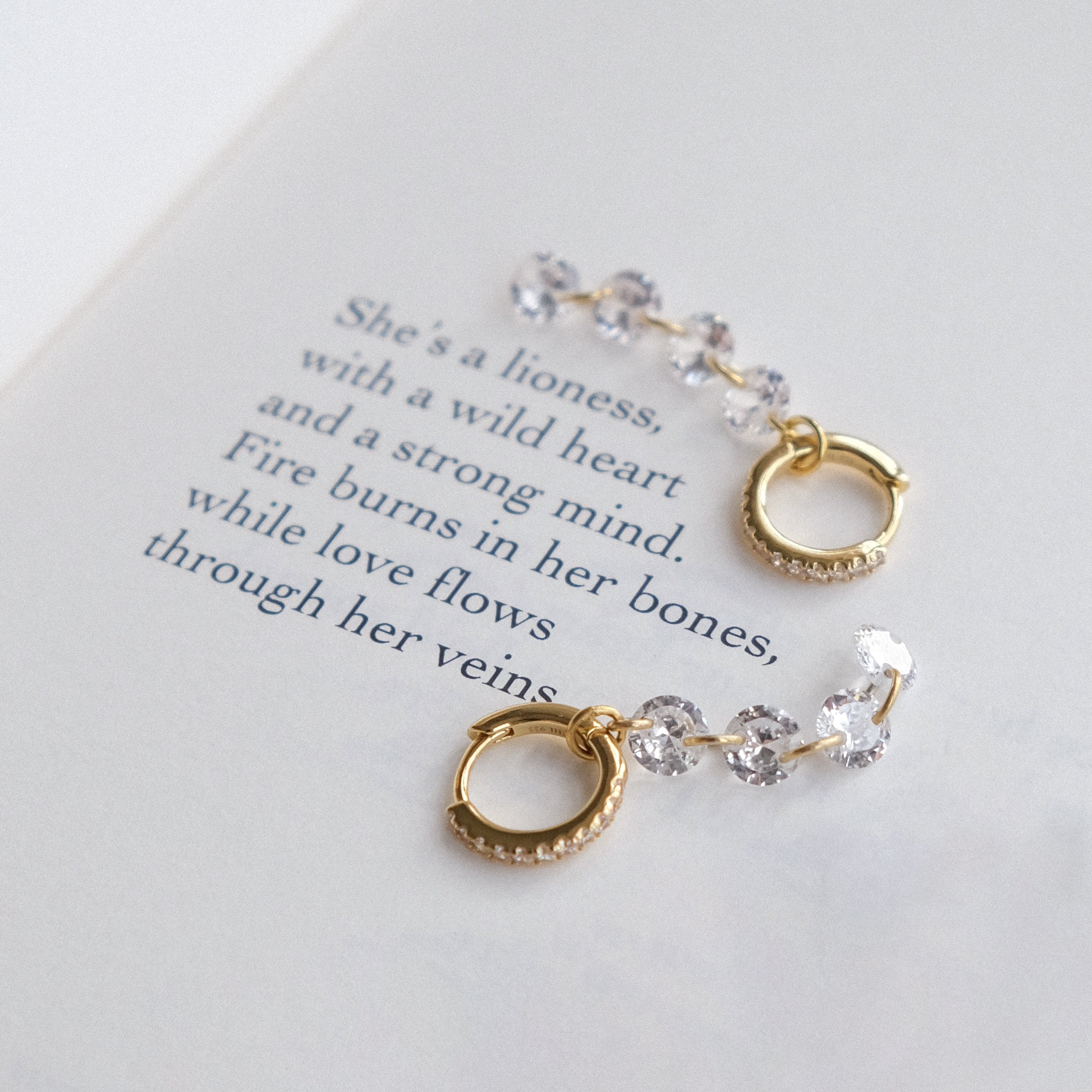 ALLURE Cascading Crystal Huggie Hoops by Crystal Heaven London, featuring clear gemstones, rest on a page with poetic text about a lioness and wild heart.