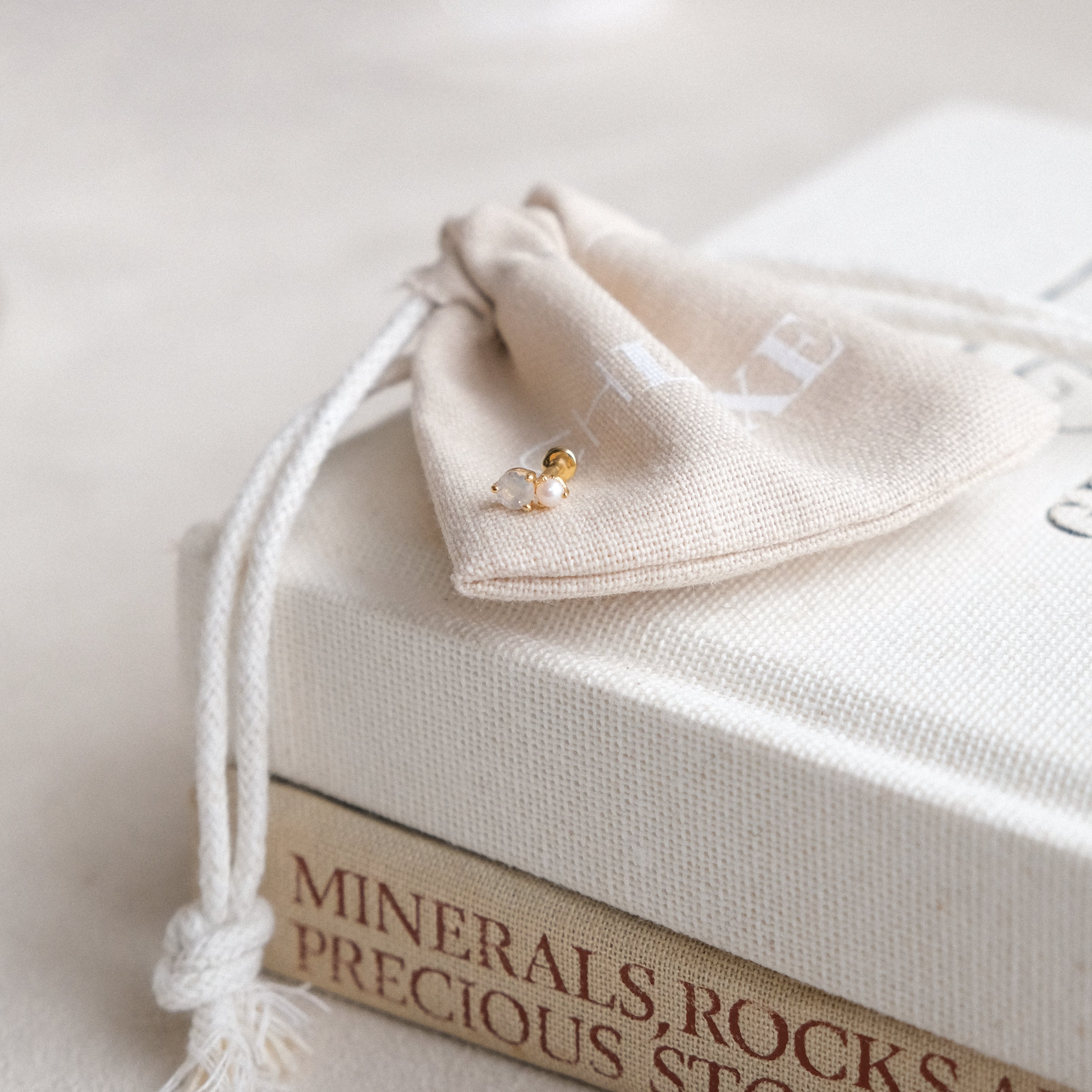 A small cloth drawstring bag sits on stacked books with a pair of pearl earrings. Nearby, the CHLUXE • OPHELIA Cartilage Stud in 14K yellow gold by Crystal Heaven London shimmers softly, adding sophistication to the serene scene.