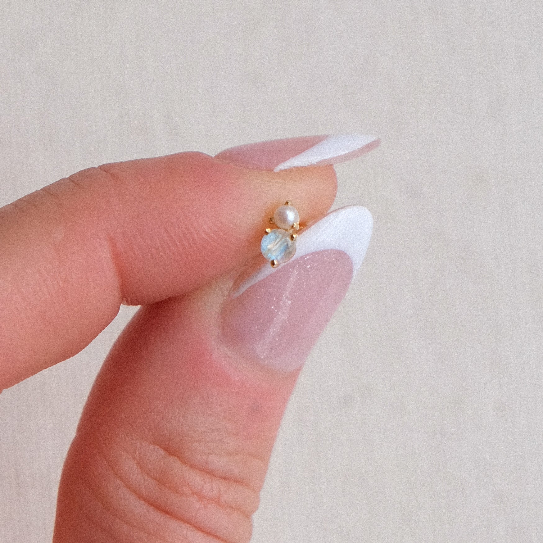 Close-up of a hand holding the CHLUXE • OPHELIA Cartilage Stud from Crystal Heaven London in 14K yellow gold, adorned with a pearl and opalescent stone. The person's nails are perfectly manicured with a French tip design.