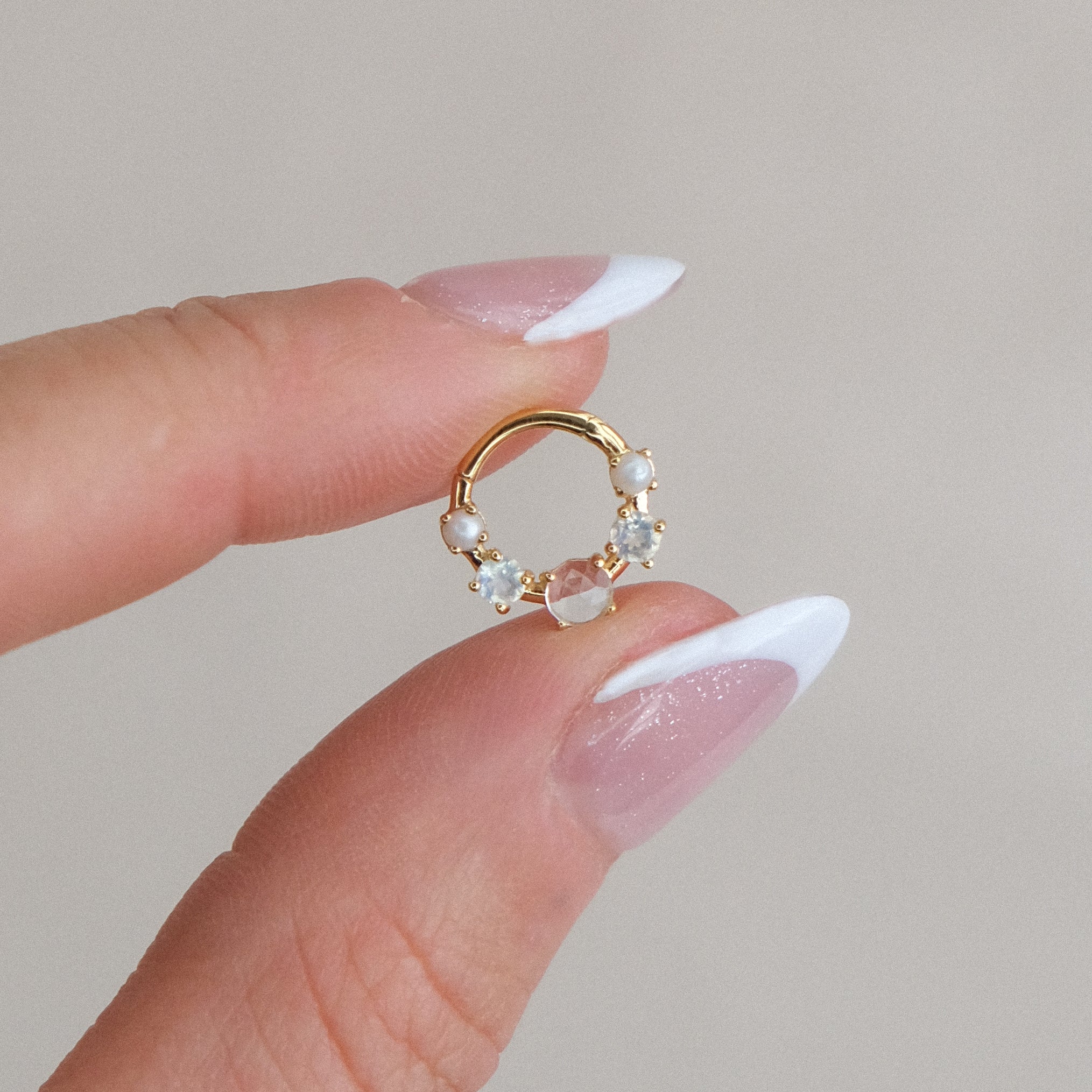 A manicured hand gently holds the CHLUXE • OPHELIA Gemstone Daith Hoop by Crystal Heaven London, crafted from 14K yellow gold and adorned with pearls and gemstones, set against a neutral background.