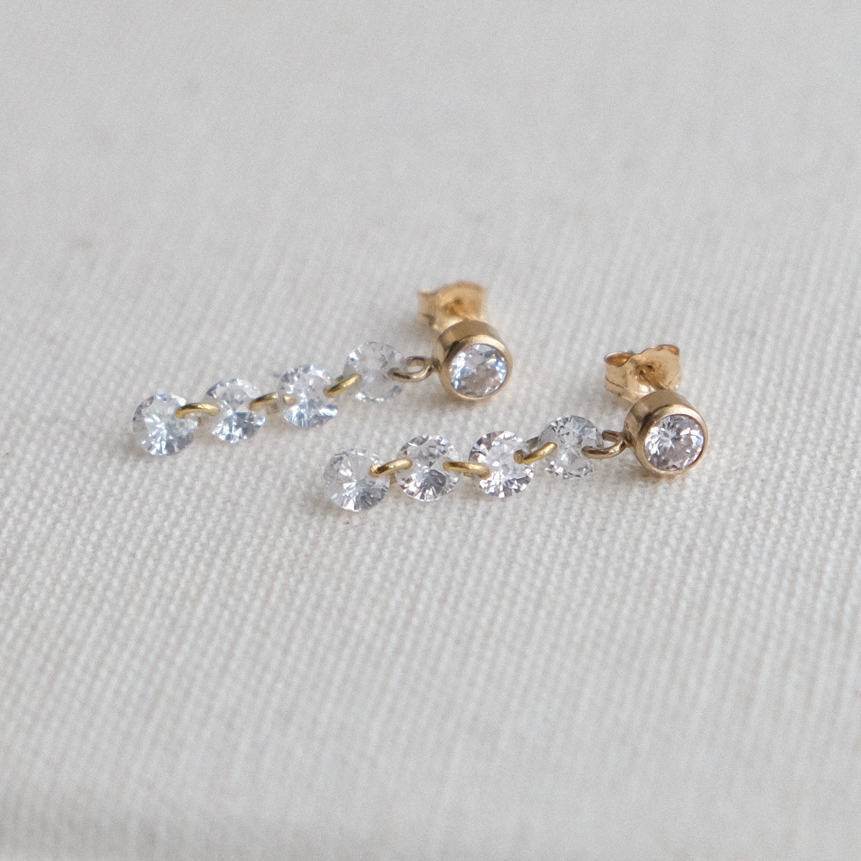 The ALLURE Cascading Crystal Studs by Crystal Heaven London feature gold studs with cascading crystals, elegantly dangling in chains of small round gemstones on a light fabric background.