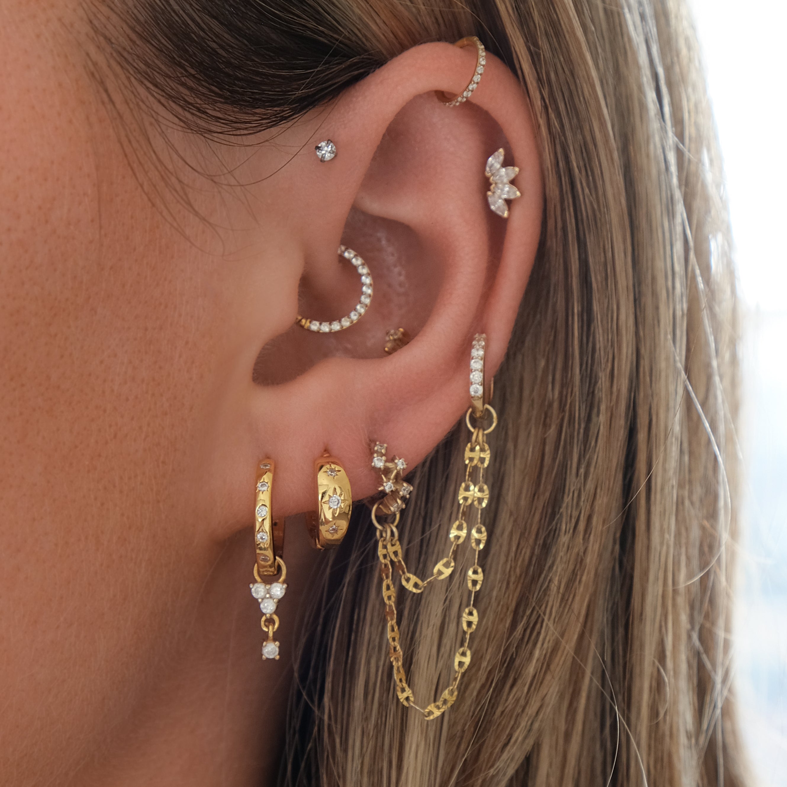An ear featuring a range of gold and diamond earrings, like hoops, studs, and the HAVANA Slim Starburst Huggie Hoop by Crystal Heaven London, adorned with chain-linked starburst details to showcase diverse piercings.