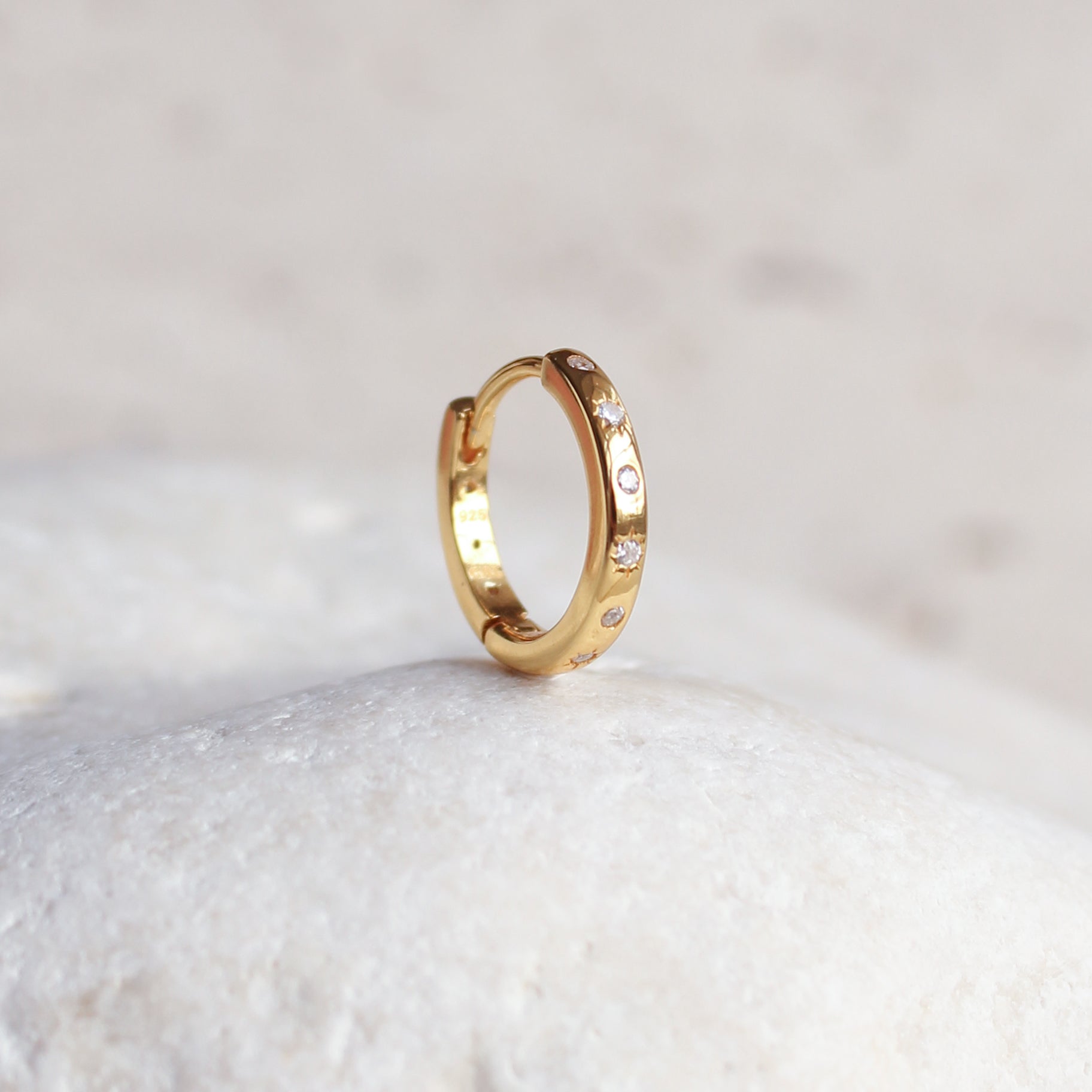 The HAVANA Slim Starburst Huggie Hoop from Crystal Heaven London, a small gold hoop earring with starburst detailing, rests on a smooth, white stone surface.