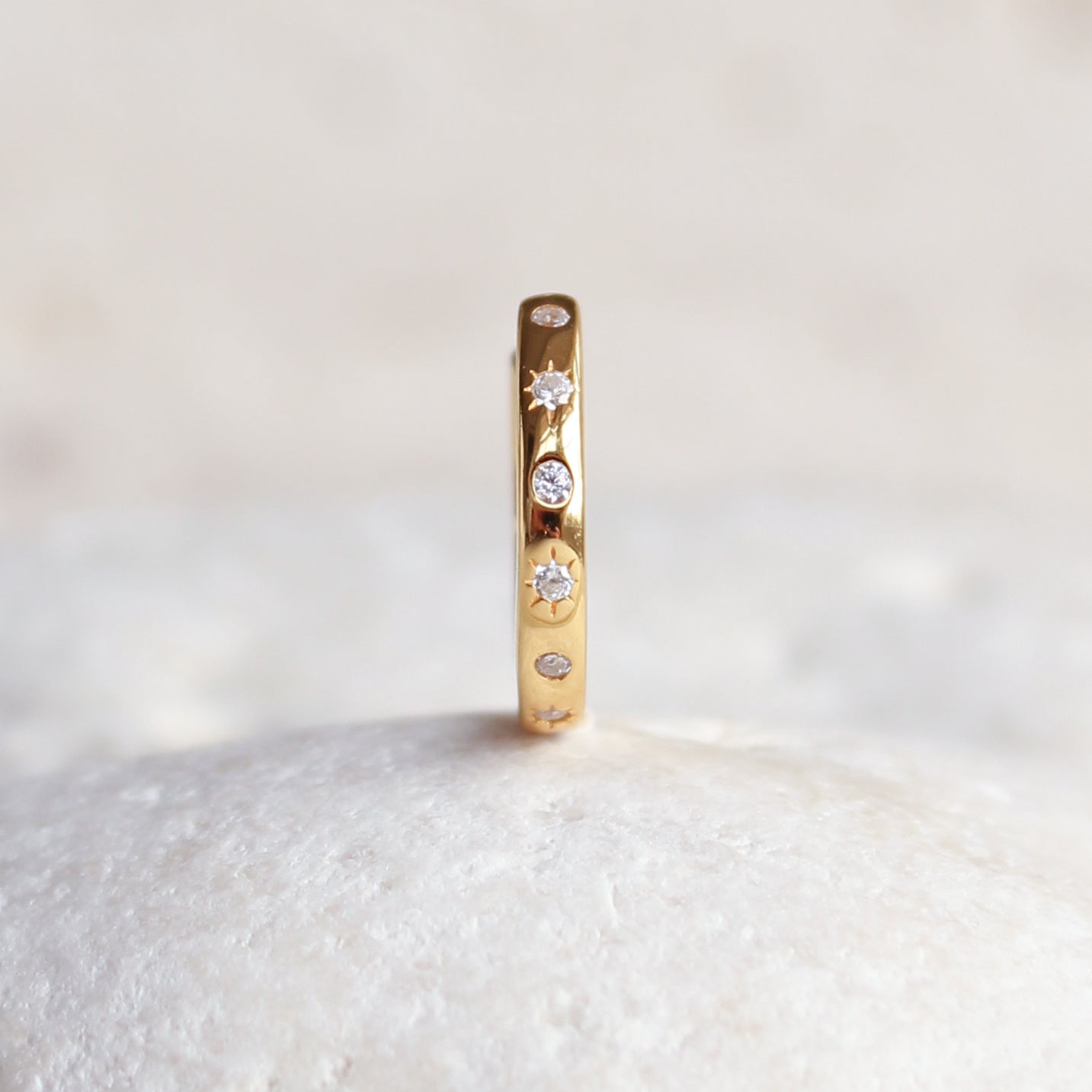 The HAVANA Slim Starburst Huggie Hoop by Crystal Heaven London, made of gold with subtle starburst detailing and small white gemstone accents, is elegantly set upright on a white textured surface.