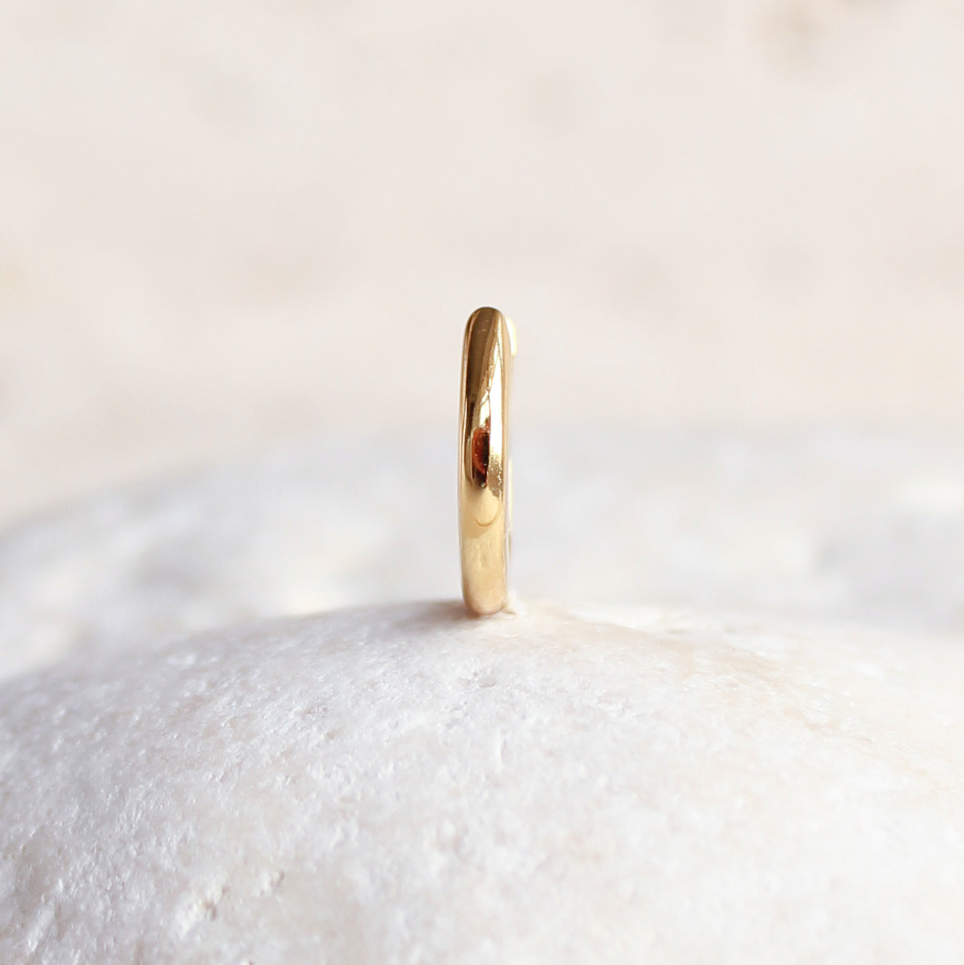 The CHLUXE Classic Huggie Hoop by Crystal Heaven London, a 14K yellow gold piece, stands upright on a textured white surface, exuding elegance akin to a timeless heirloom earring.