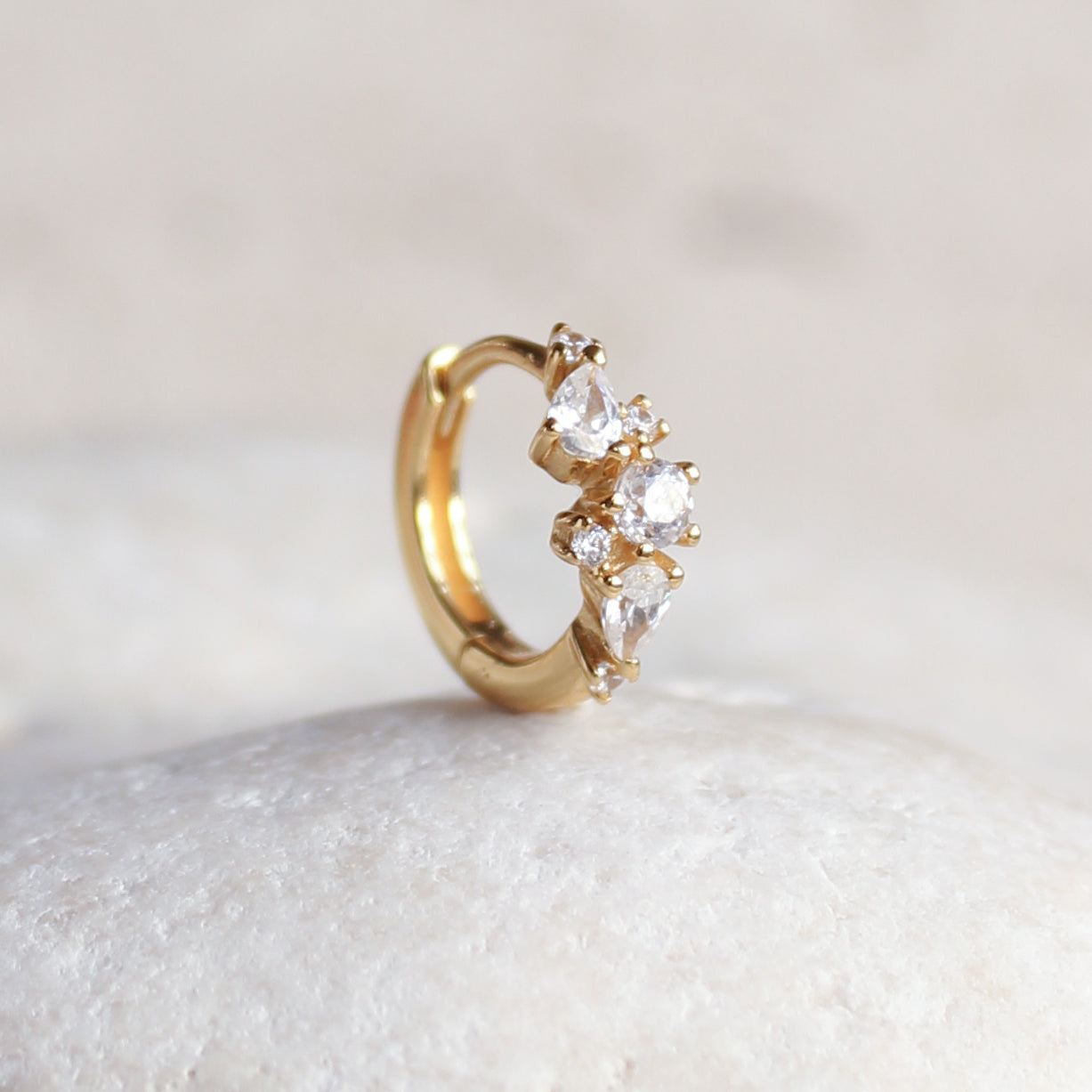 The MARGOT Scattered Gem Huggie Hoops by Crystal Heaven London, adorned with five clear cubic zirconia stones, are displayed on a smooth, white stone surface.