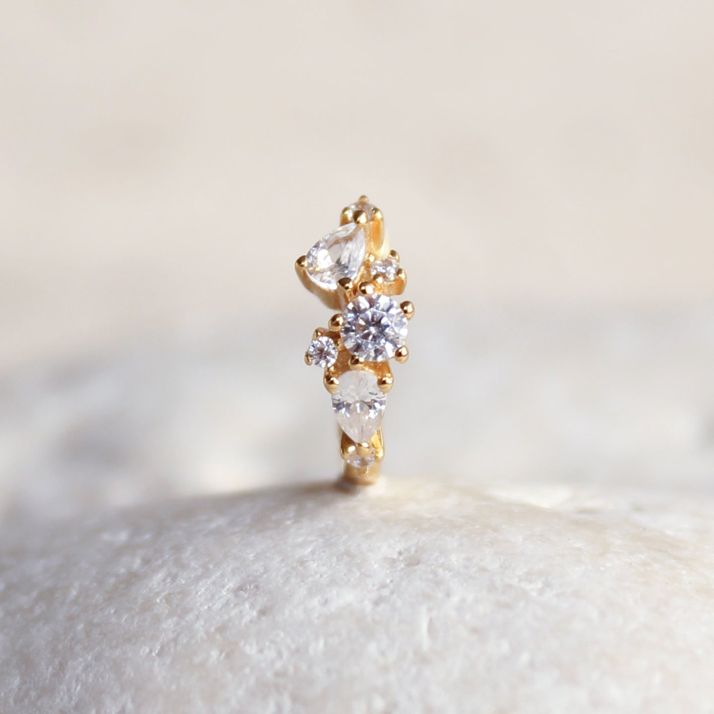 The MARGOT Scattered Gem Huggie Hoops by Crystal Heaven London feature an asymmetric design with round and teardrop-shaped cubic zirconia, elegantly displayed on a smooth white surface.