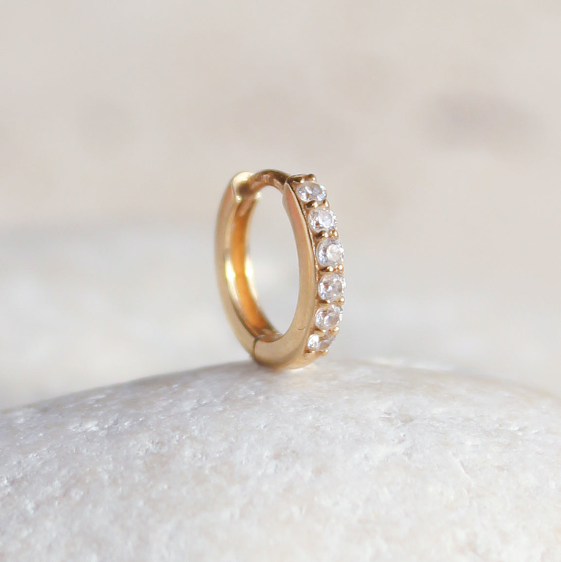 The CHLUXE Crystal Huggie Hoop by Crystal Heaven London, crafted in 14K yellow gold and adorned with clear gemstones, elegantly rests on a smooth white surface.
