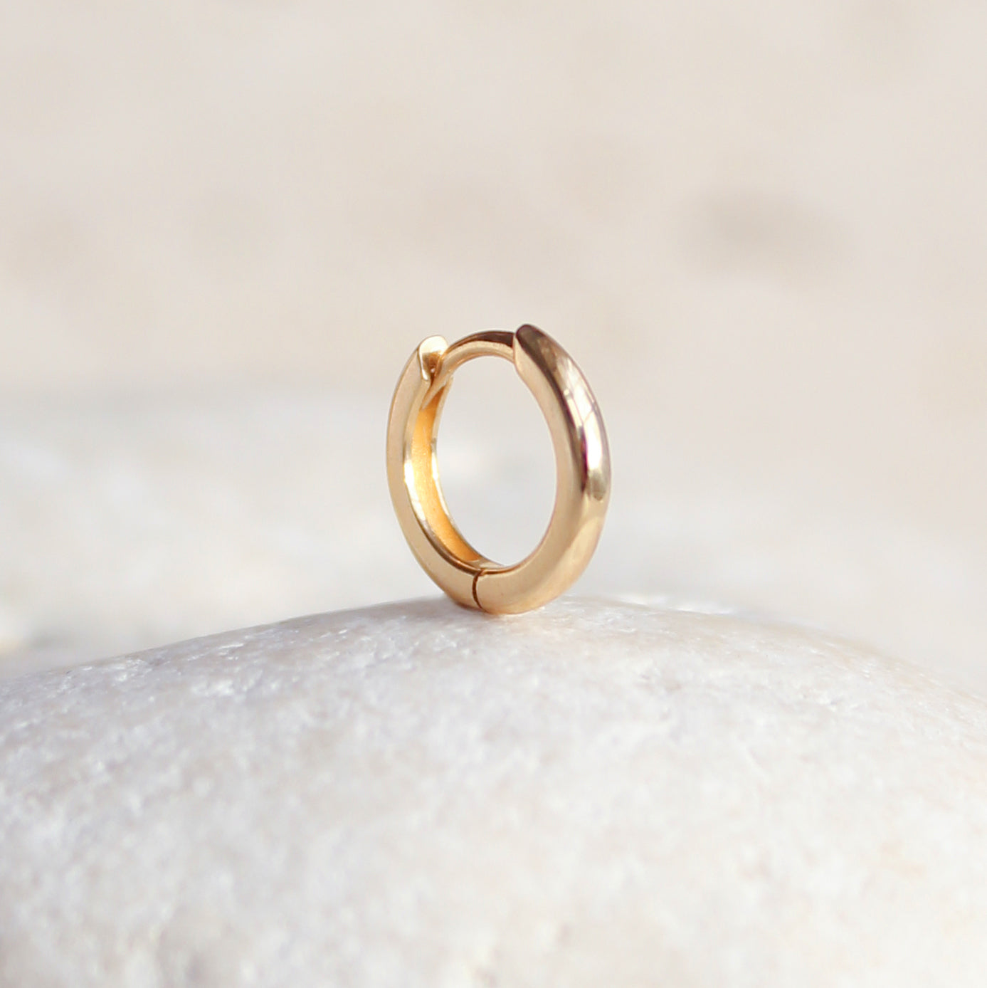 The CHLUXE Classic Huggie Hoop by Crystal Heaven London, crafted in 14K yellow gold, elegantly rests upright on a smooth white stone surface.