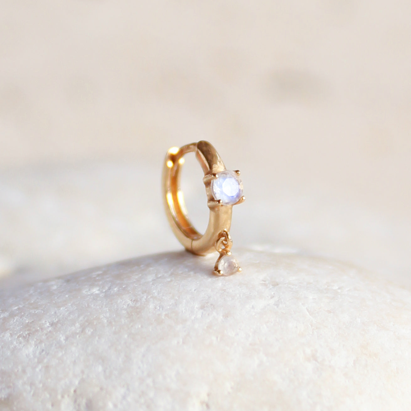 The CHLUXE Moonstone Droplet Huggie Hoop by Crystal Heaven London, featuring a round white moonstone and teardrop charm in 14K yellow gold, rests elegantly on a smooth white surface.