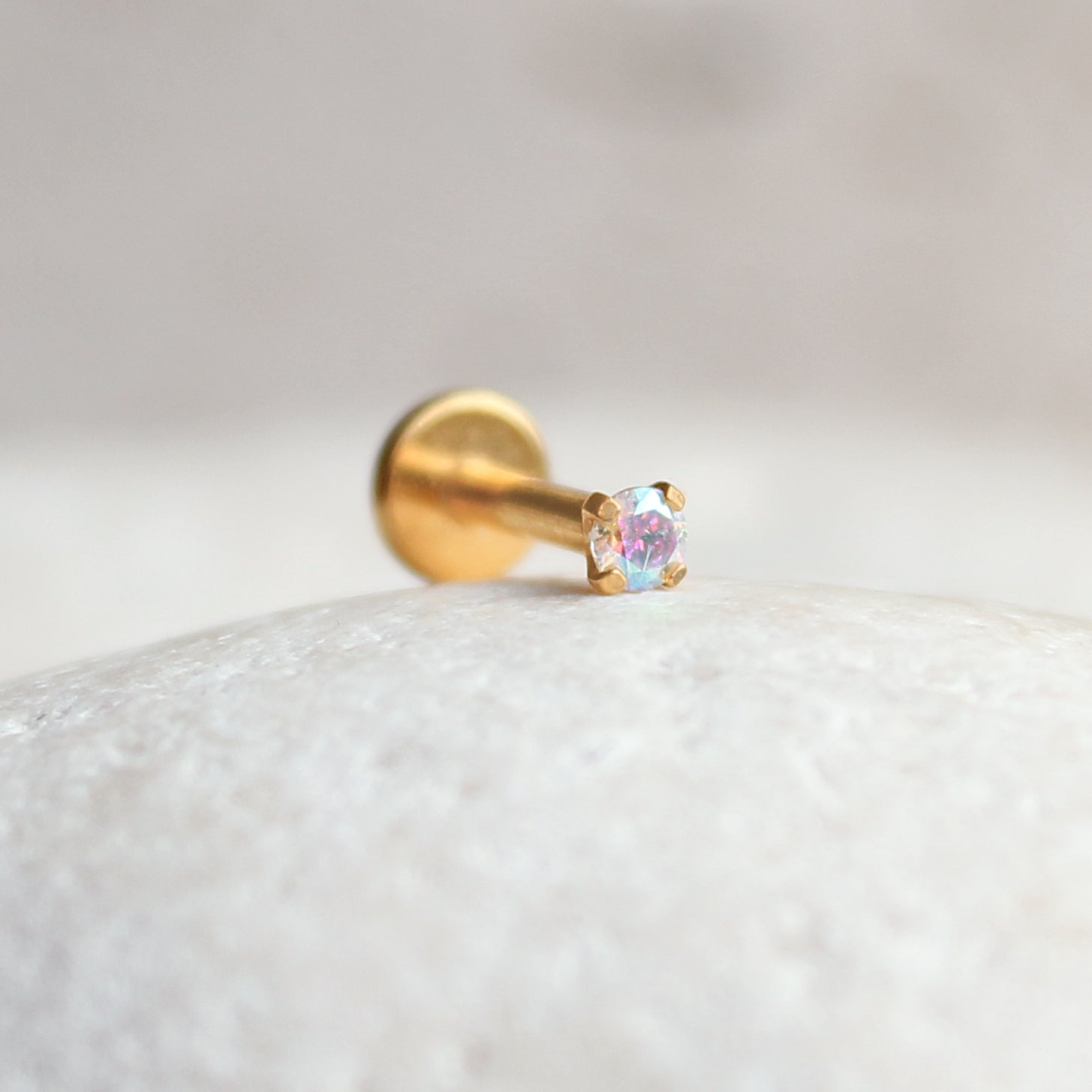 The Crystal Heaven London Aurora Rainbow Solitaire Cartilage Stud, a gold stud earring featuring a shimmering crystal, rests elegantly on a textured, light-colored surface, ideal for your ear stack.