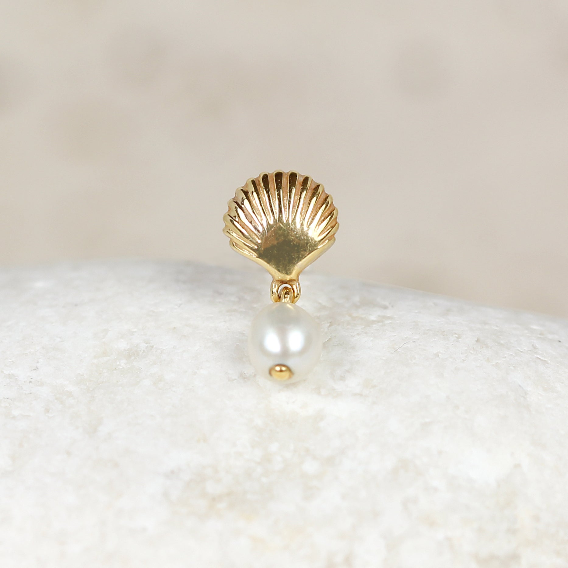The CHLUXE • MILOS Cartilage Stud, a limited edition 14k Yellow Gold earring by Crystal Heaven London, elegantly features a shell shape with a dangling white pearl resting on a smooth, light-colored stone surface.