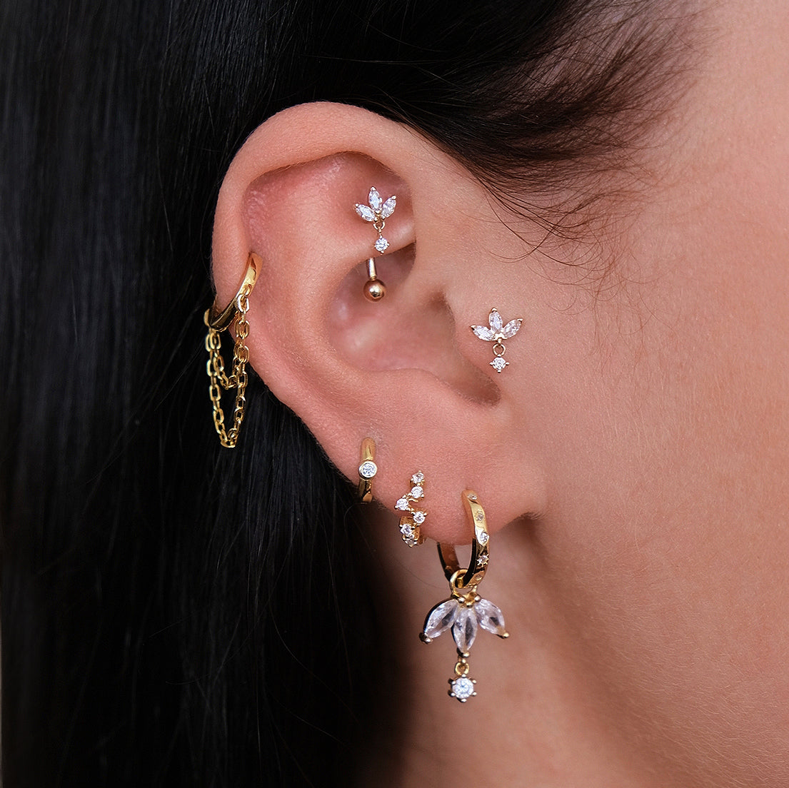 A close-up showcases an ear adorned with 14k yellow gold and diamond earrings, featuring hoops, chains, and floral designs. The rook piercing is elegantly highlighted by the Crystal Heaven London CHLUXE LANI Droplet Curved Rook Barbell.