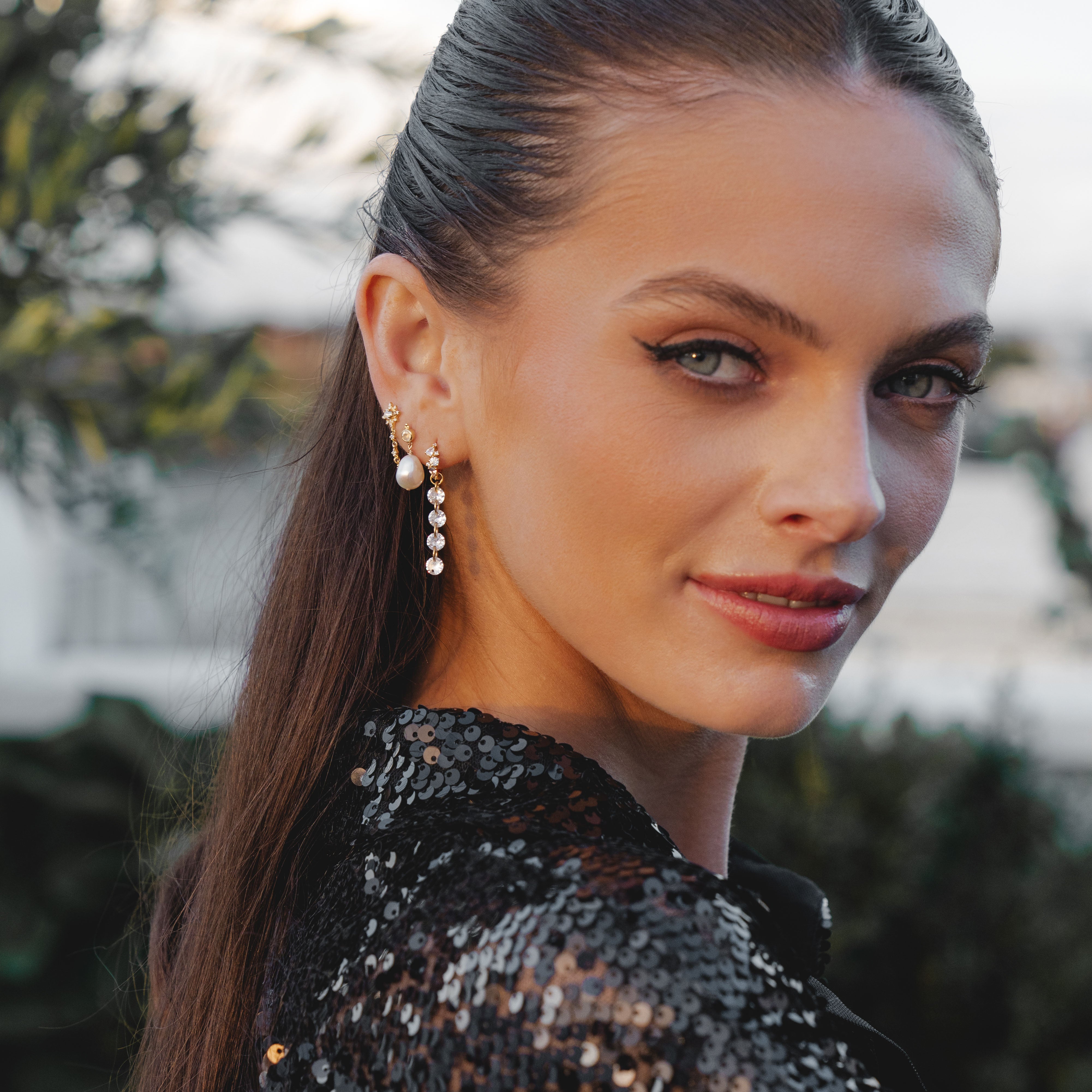 With long dark hair and wearing a sequined black garment, someone exudes confidence in the ALLURE Cascading Crystal Earring Charm by Crystal Heaven London. The lush greenery backdrop enhances their allure.