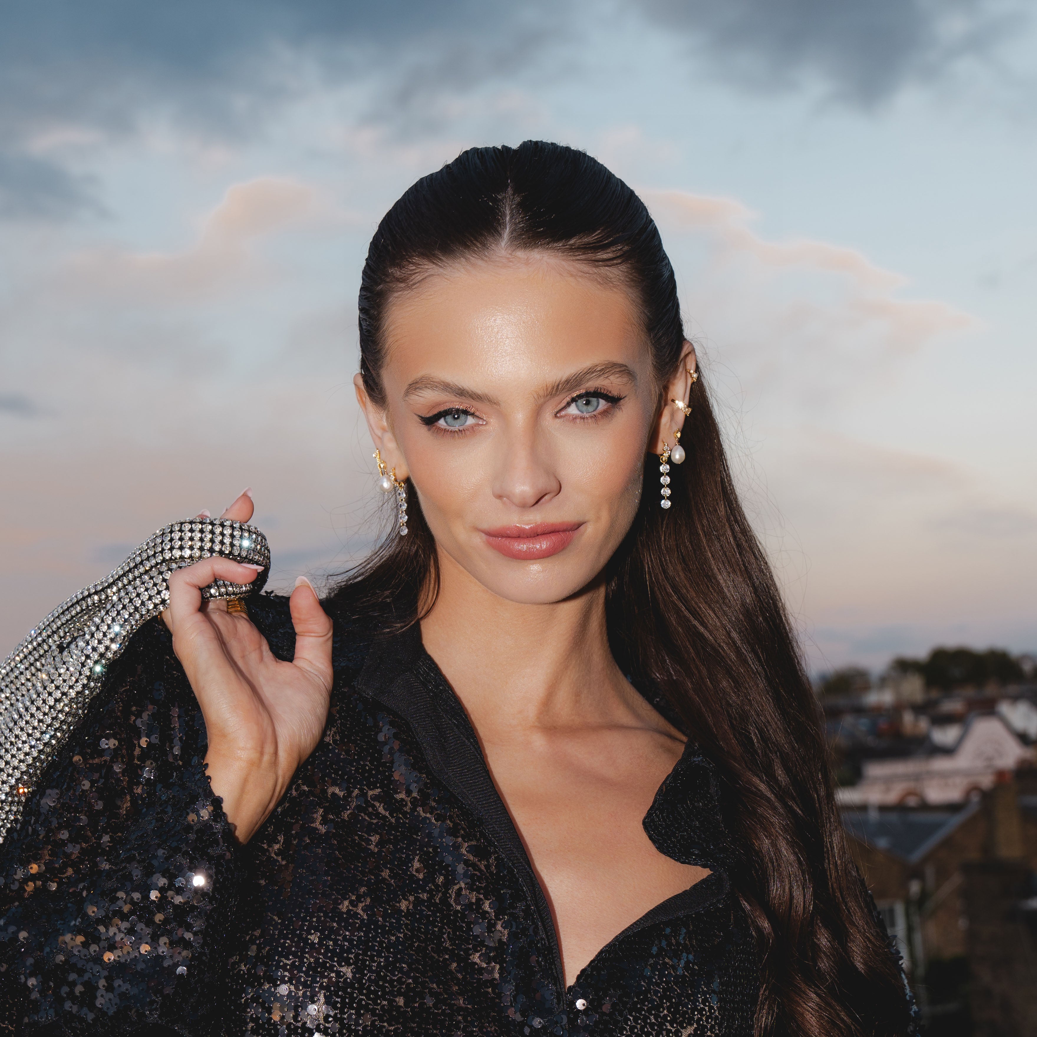 A woman with long dark hair in a sequined outfit poses outdoors, holding shimmering ALLURE Cascading Crystal Huggie Hoops by Crystal Heaven London over her shoulder against a dusky sky.