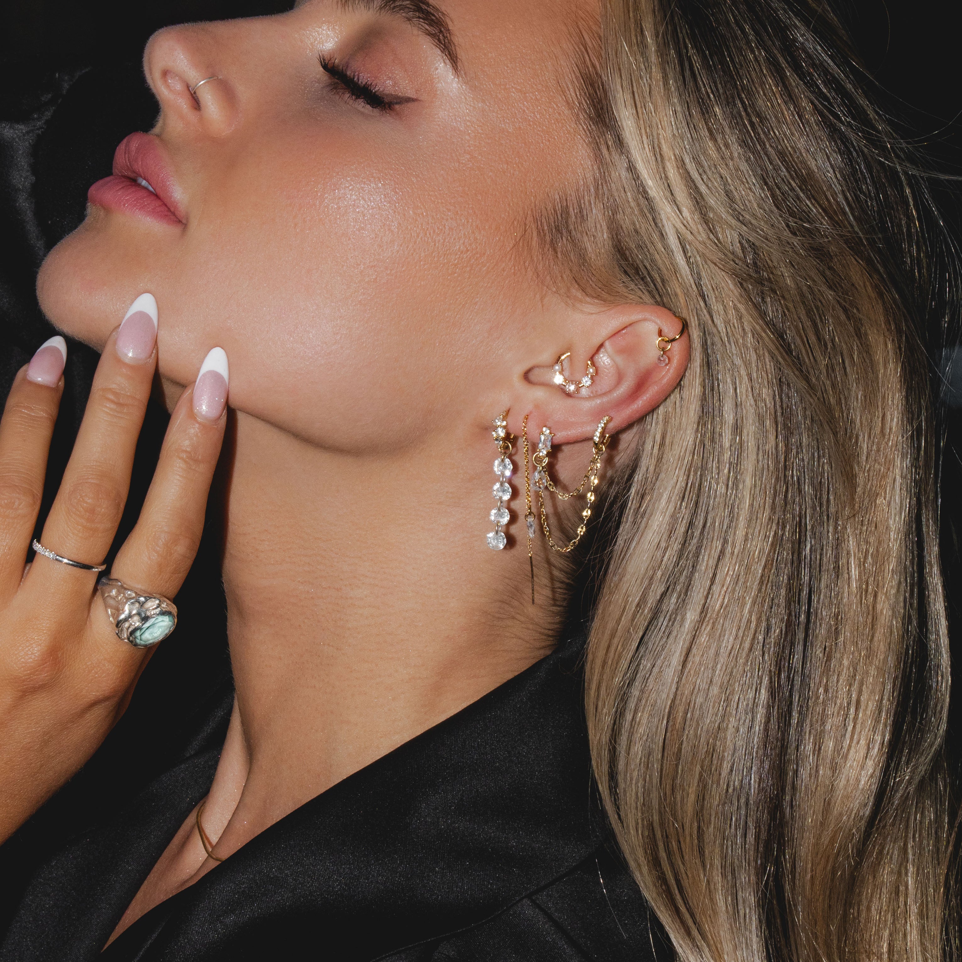A woman with long blonde hair, elegantly wearing the CHLUXE • OPHELIA Gemstone Daith Hoop from Crystal Heaven London in 14K yellow gold, dressed in black, gently touching her face with a manicured hand—a modern Ophelia.