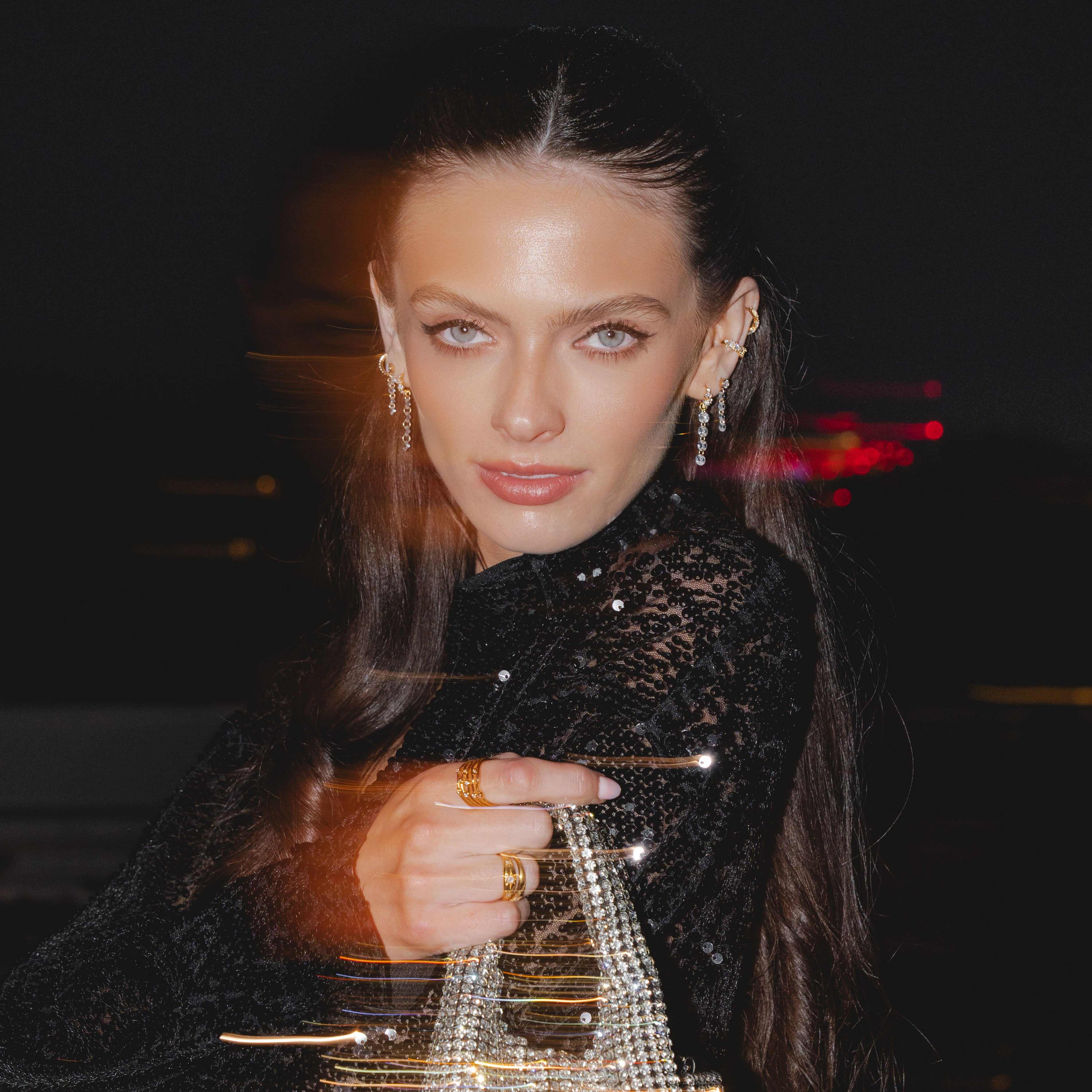 A woman with long hair poses holding a sparkling handbag against a dark backdrop, wearing a black outfit and captivating ALLURE Cascading Crystal Studs by Crystal Heaven London that glisten beautifully.