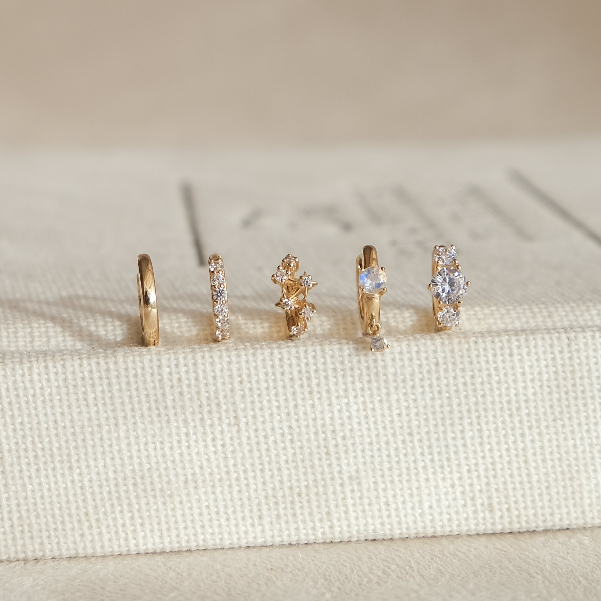 Five CHLUXE Moonstone Droplet Huggie Hoops by Crystal Heaven London, crafted in 14K yellow gold and adorned with various gemstones, stand upright on a textured beige surface.