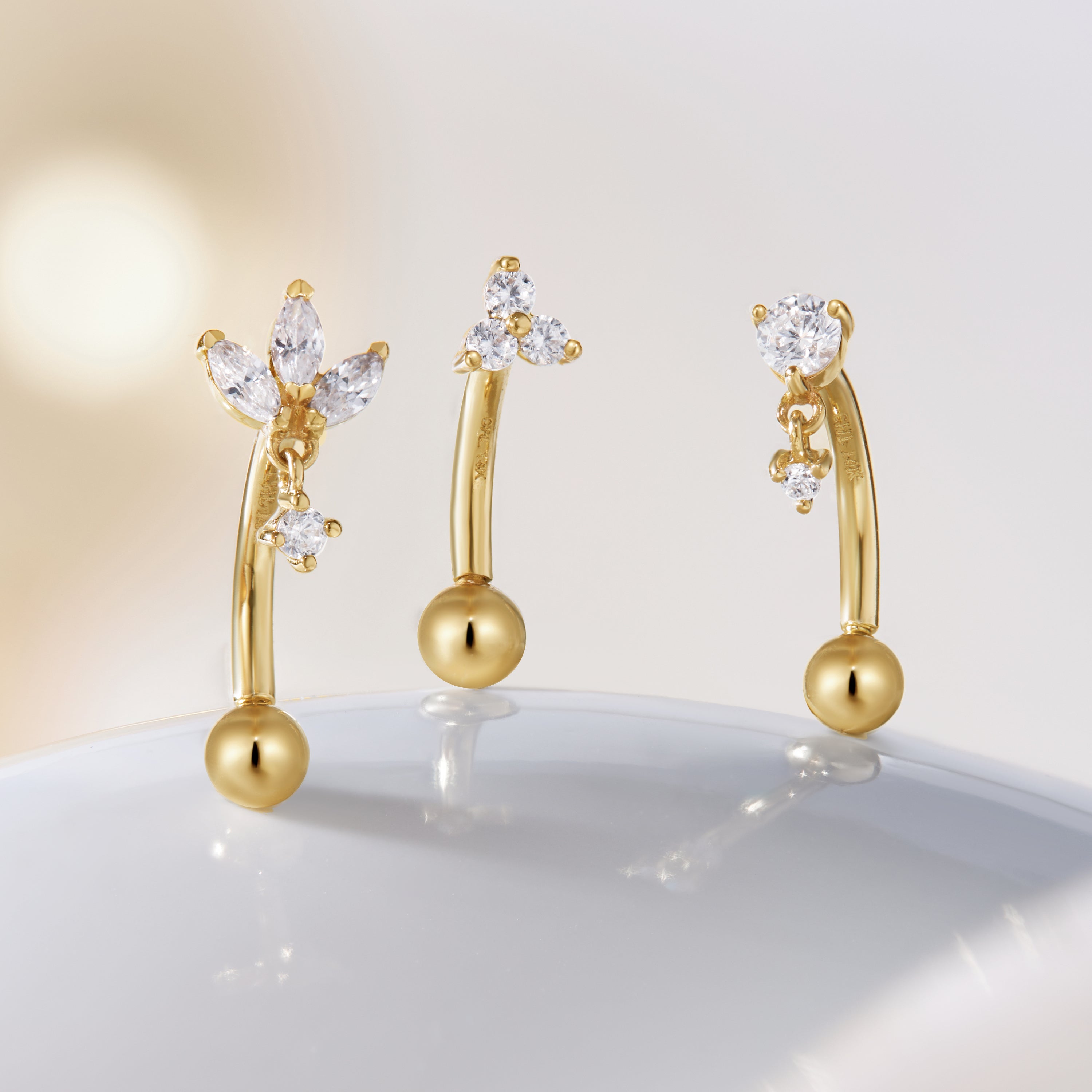 On a smooth white surface against a soft, blurred background are three CHLUXE LANI Droplet Curved Rook Barbells in 14K Yellow Gold from Crystal Heaven London, each featuring diamond accents that add elegance to their gold curves.