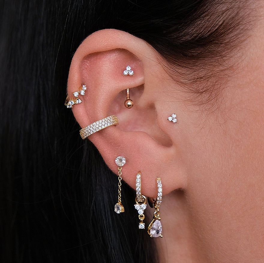 Close-up of an ear with multiple piercings, including a rook piercing featuring the CHLUXE Trinity Curved Rook Barbell in 14K white gold by Crystal Heaven London. The earrings, from studs to hoops and chains, create a striking contrast with their blend of gold tones.