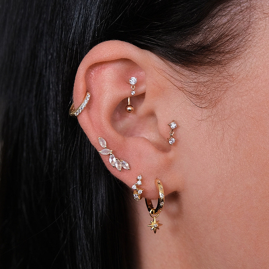 A close-up of an ear showcases several gold and diamond earrings, including hoops, studs, a dangling piece, and an elegant Crystal Heaven London CHLUXE Vogue Curved Rook Barbell in 14k Yellow Gold.