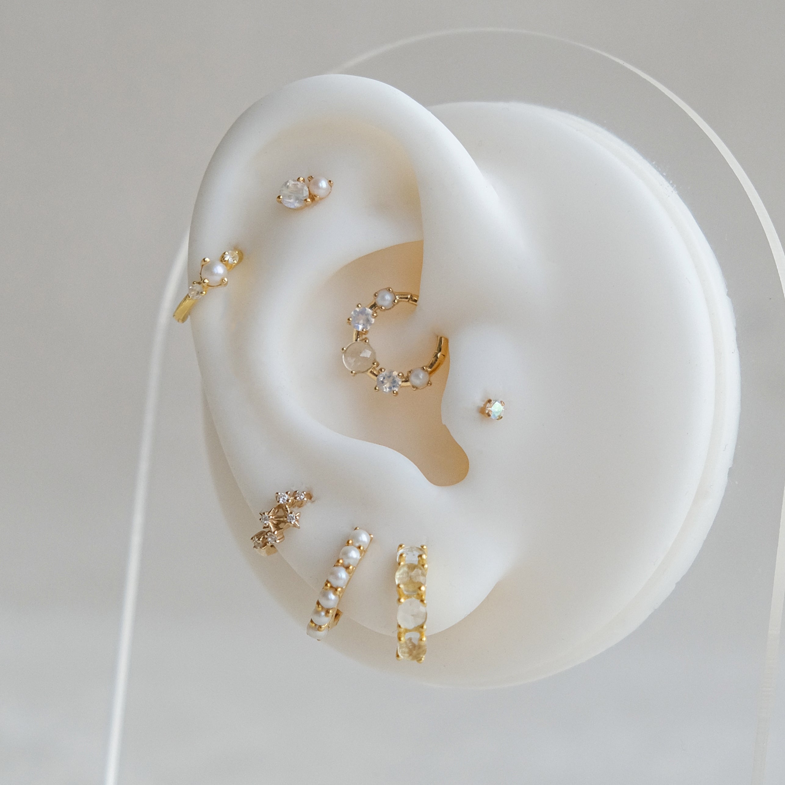 Crystal Heaven London's Aurora Rainbow Solitaire Cartilage Stud and other gold, pearl, and aurora crystal earrings decorate a white ear model, showcasing varied styles for the ideal ear stack. Made with hypoallergenic titanium, each piece balances comfort and elegance.