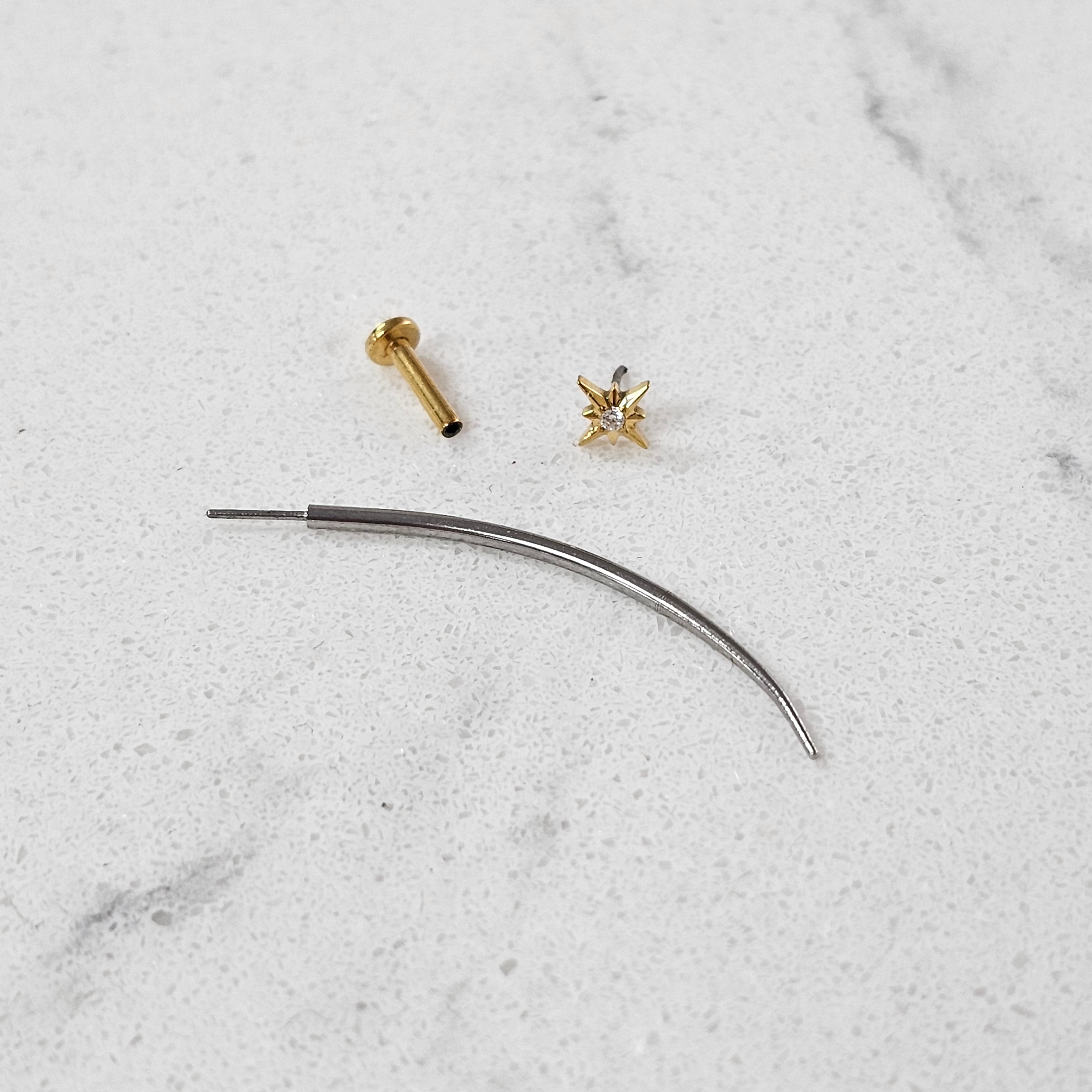 A sleek Crystal Heaven London Curved Labret Insertion Tool (Threadless) made of 316L stainless steel, a small gold stud, and a charming gold star rest elegantly on the white surface.