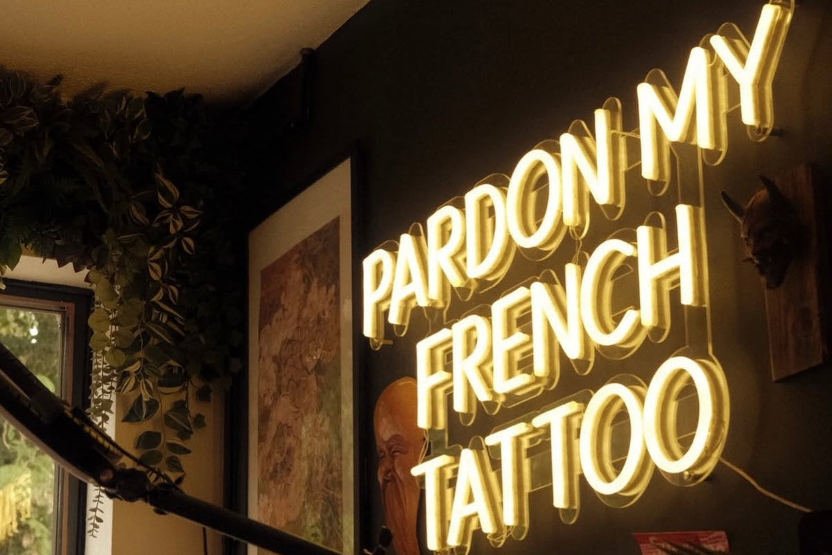 A glowing neon sign in a tattoo studio reads 'PARDON MY FRENCH TATTOO' in bright yellow letters against a dark wall, with framed artwork and tattoo supplies visible around it.