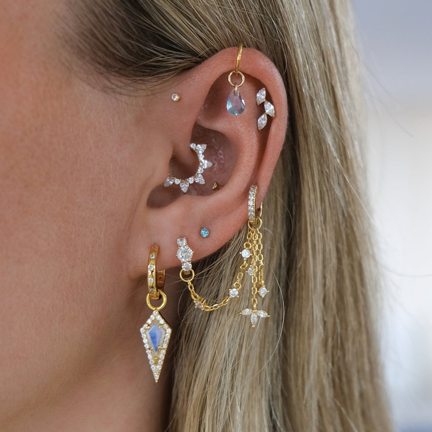 Close-up of an ear adorned with multiple earrings, including the AURA Mini Moonstone Pavé Kite Charm by Crystal Heaven London, featuring gold, pavé crystal, and diamond hoops, studs, and dangles.