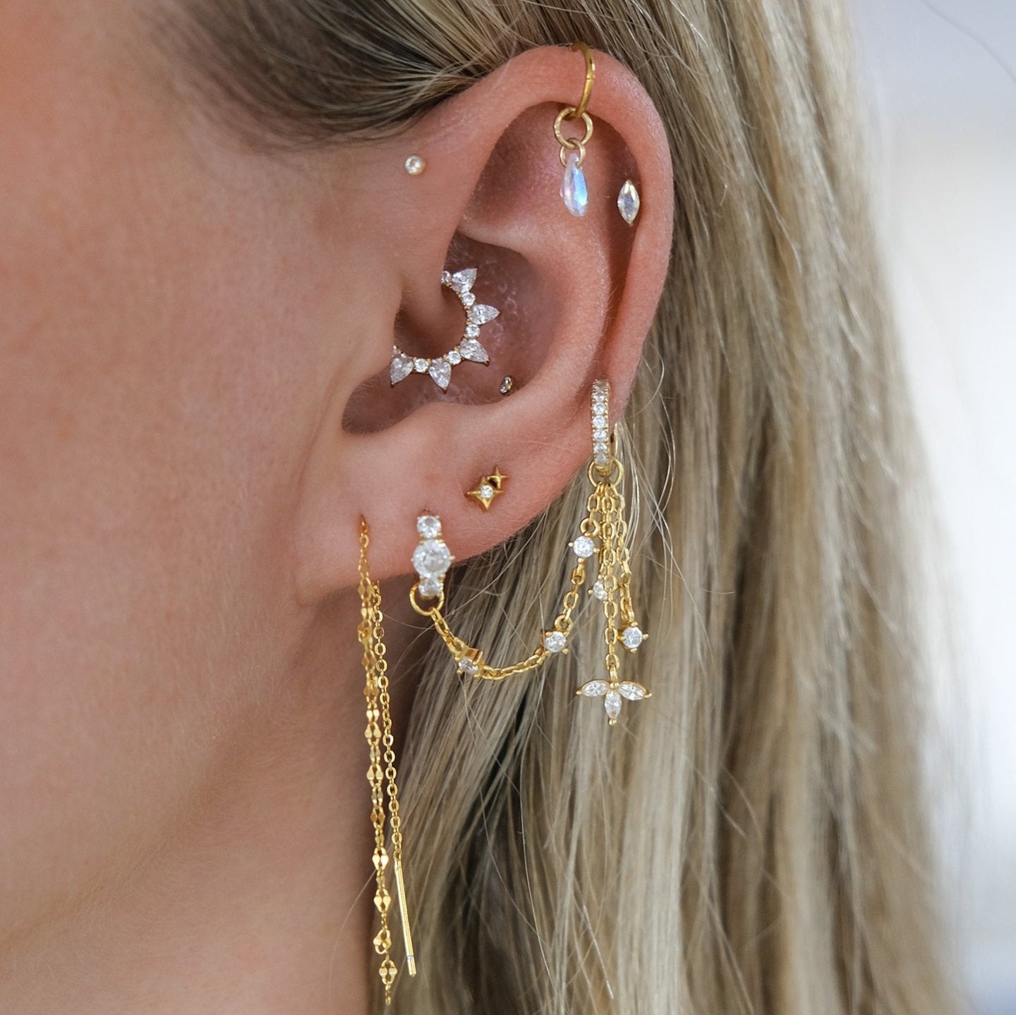 A close-up of an ear adorned with gold chains, diamond-studded designs, and teardrop crystals features the GISELLE Daith Hoop by Crystal Heaven London. The array includes various piercings on the lobe and cartilage, featuring hypoallergenic titanium craftsmanship.