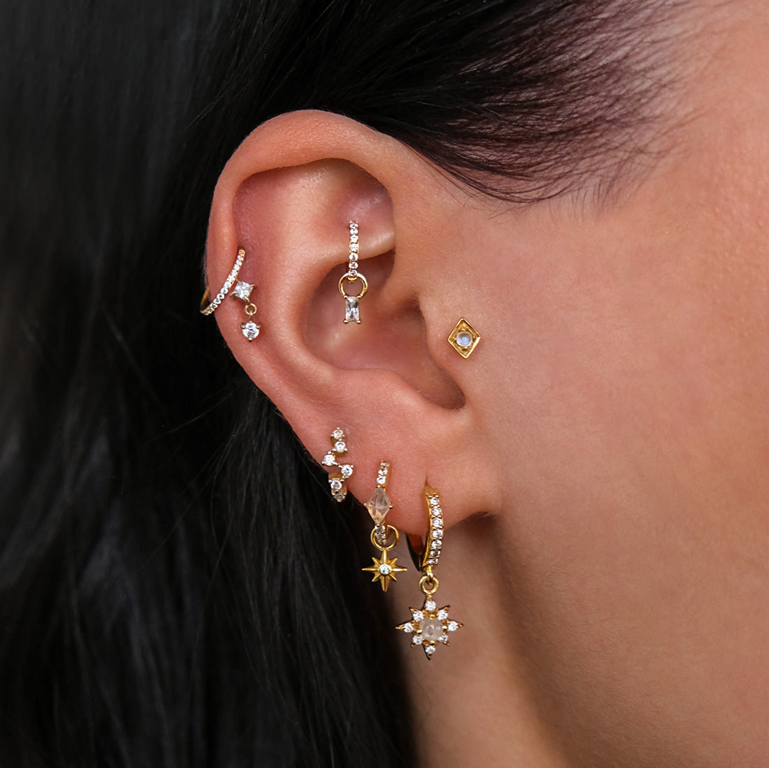 An ear adorned with multiple piercings features a stunning array of Crystal Heaven London's CHLUXE 14K Yellow Gold earrings, including the HERA Cartilage Stud. The hoops, studs, and dangly designs elegantly contrast against dark hair, adding celestial charm.